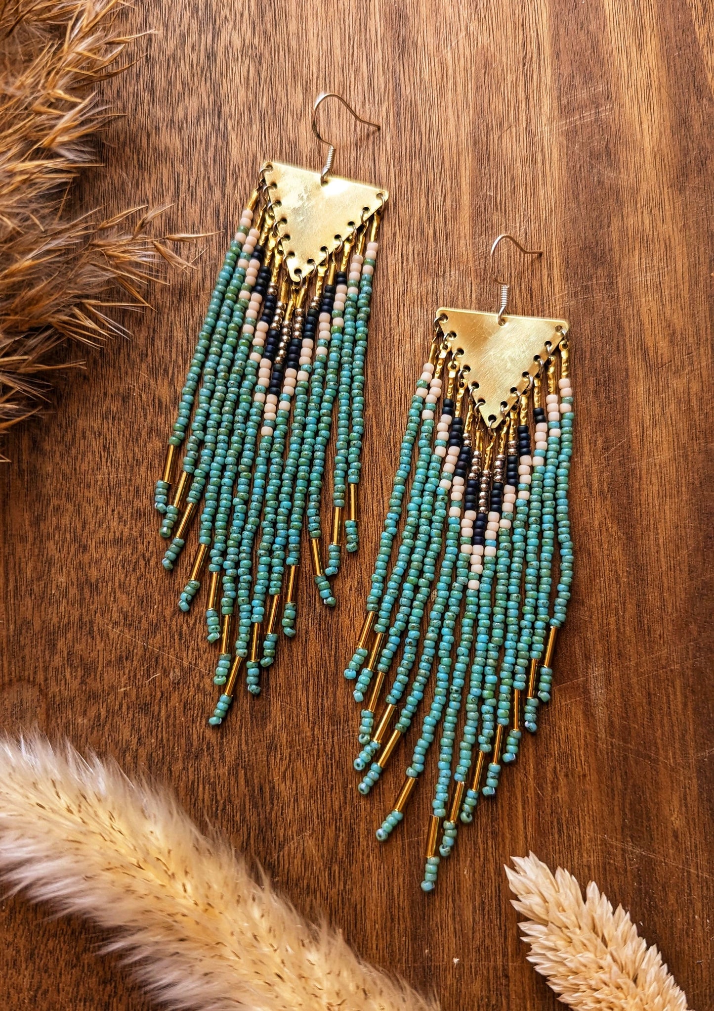 Moon & Milk - Long dangling seed bead earrings with turquoise, blue, and beige beads in a detailed geometric pattern. Gold triangular brass tops. Displayed on a wooden surface with dried grasses.