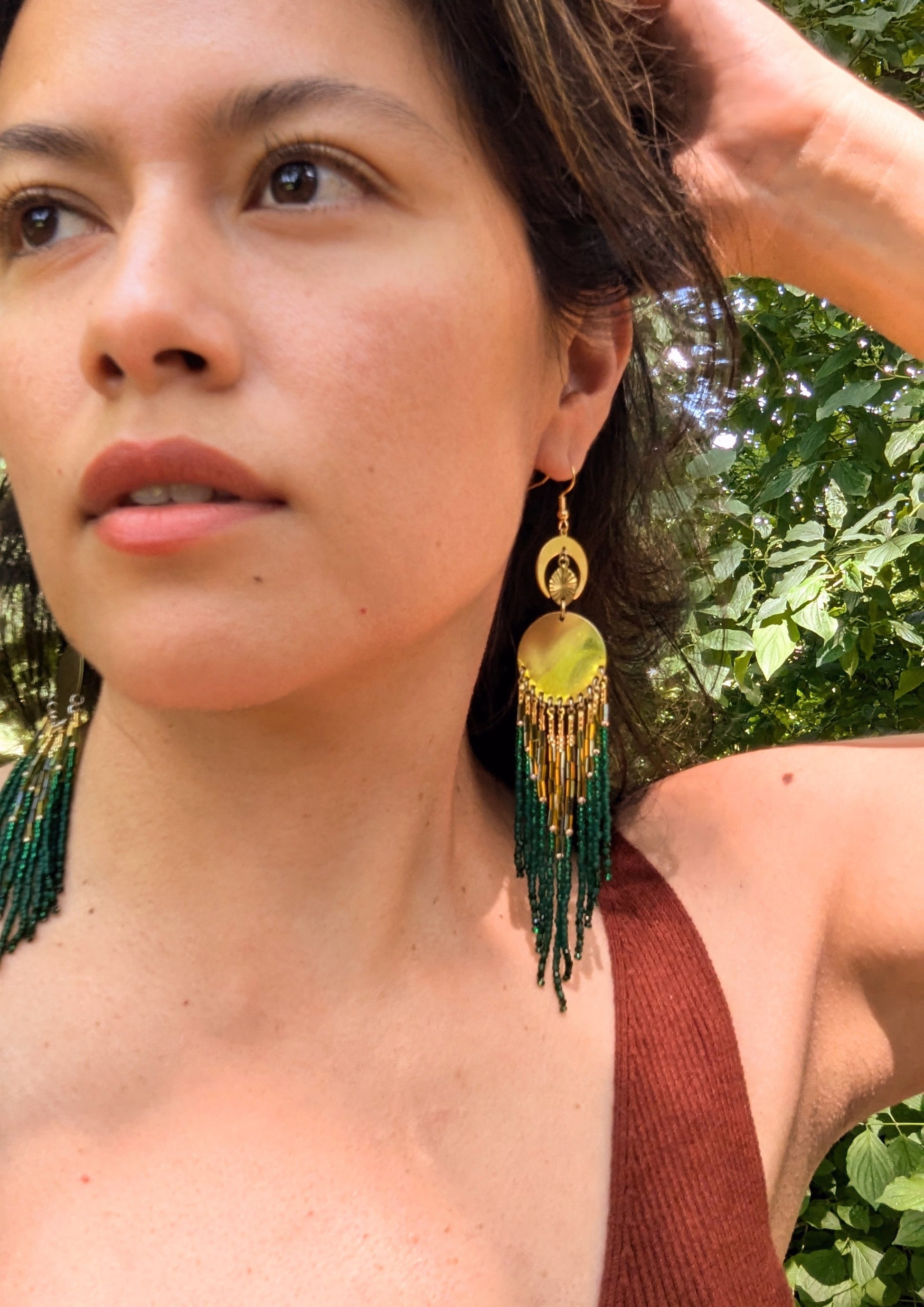 Model wearing brass and green beaded statement earrings with a crescent moon and circular design, standing out against a natural, outdoor background.- Moon & Milk