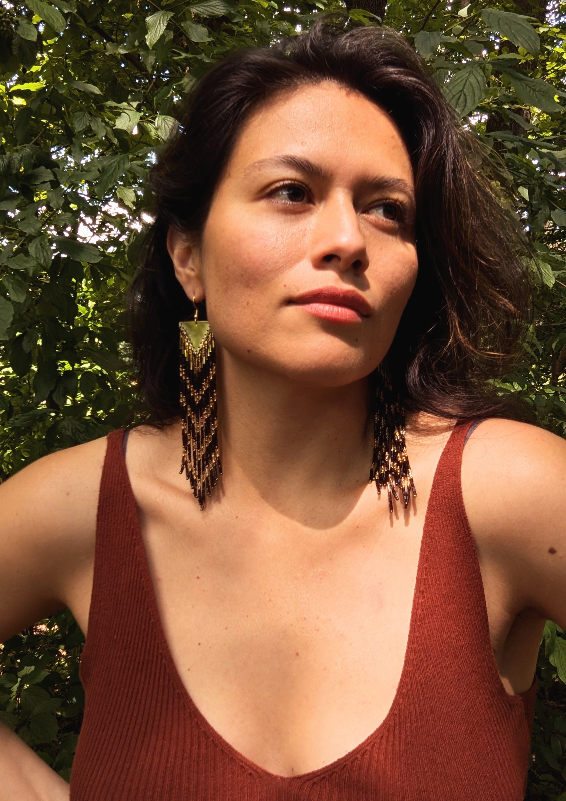 Woman wearing long native beaded earrings with dark brown and gold beads by Moon & Milk, photographed against green foliage.
