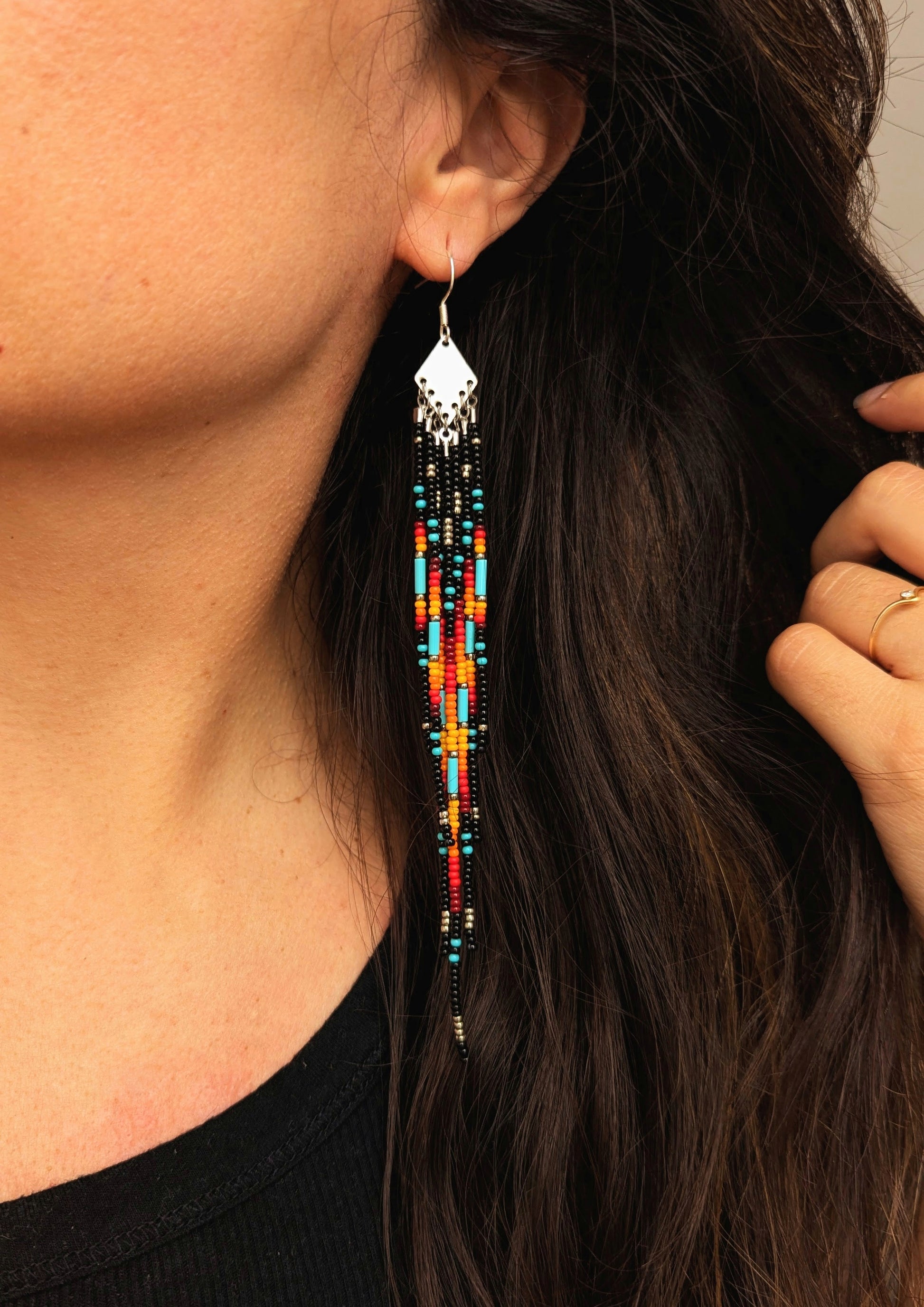Handcrafted beaded earrings on a model with a vibrant fringe design. The beads feature a bold mix of black, red, yellow, turquoise, and metallic accents, creating a striking, colorful pattern.