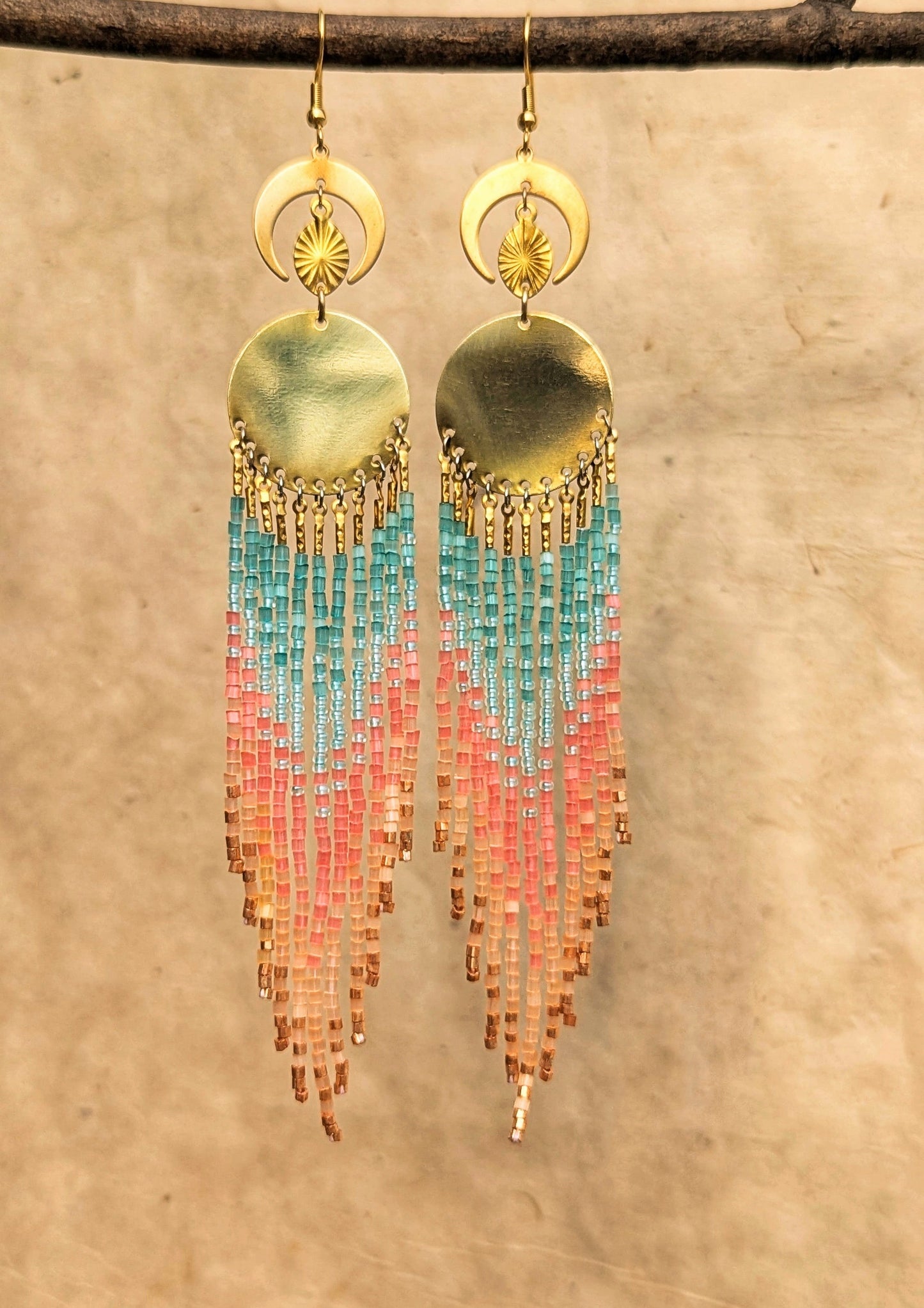 Beaded fringe earrings with crescent and sun motifs, featuring cascading beads in soft pastel shades of blue, pink, and peach, hanging against a textured background.- Moon & Milk