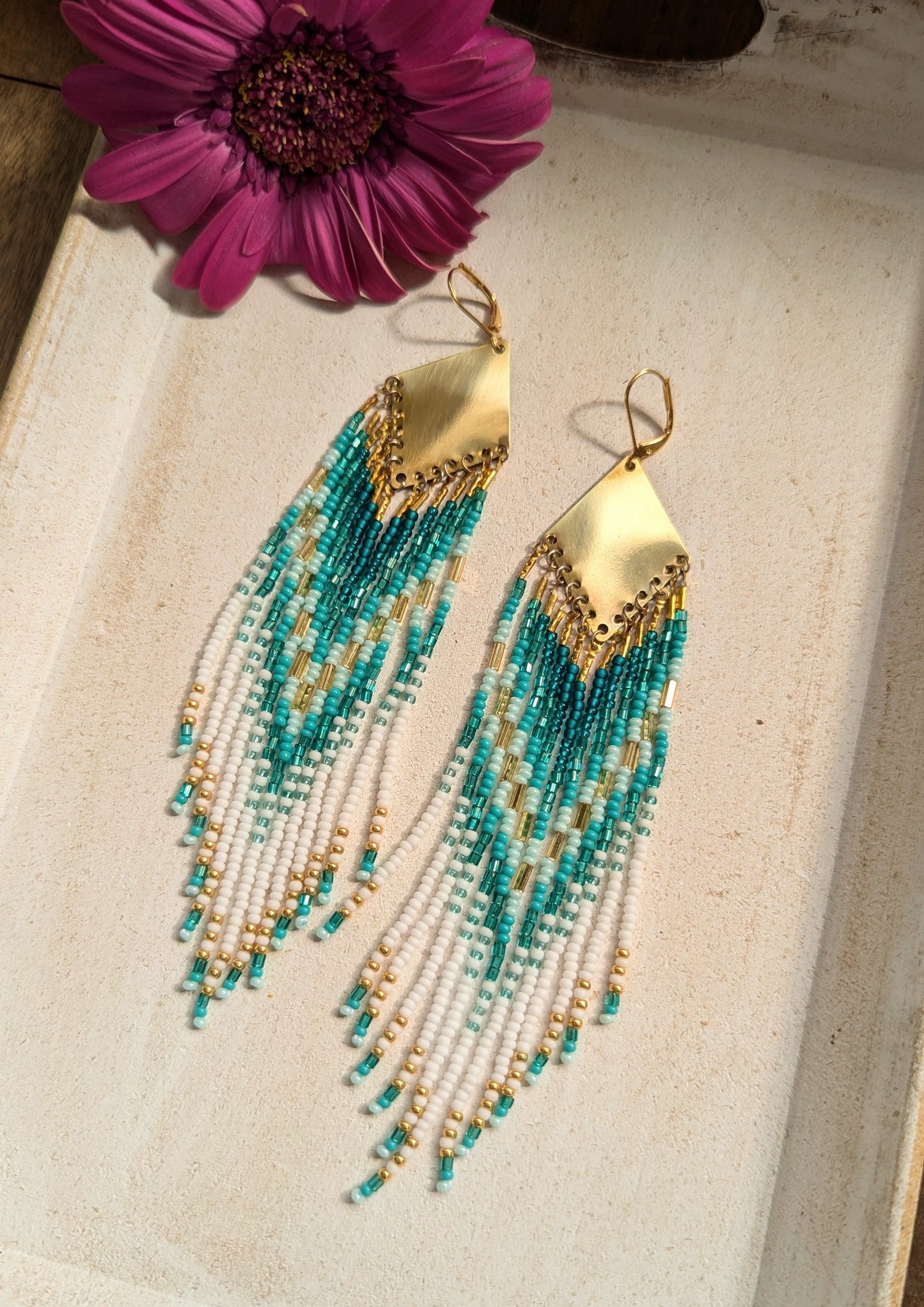 Moon & Milk turquoise, white, and gold seed bead earrings with brass findings, laid on white wood with a coral flower—hand-beaded and coastal-inspired.