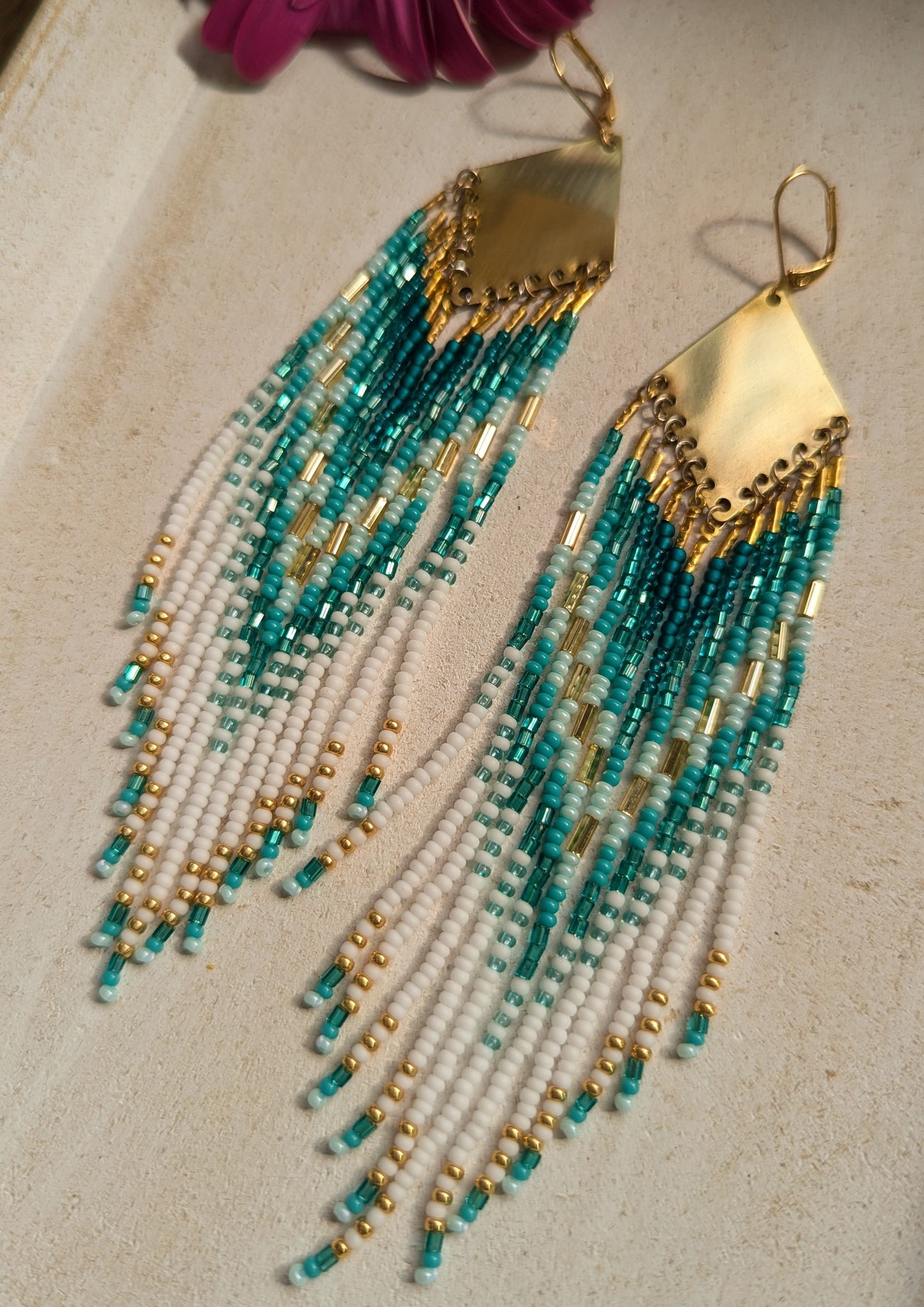 Moon & Milk turquoise, white, and gold seed bead earrings with brass findings, laid on white wood with a coral flower—hand-beaded and coastal-inspired.