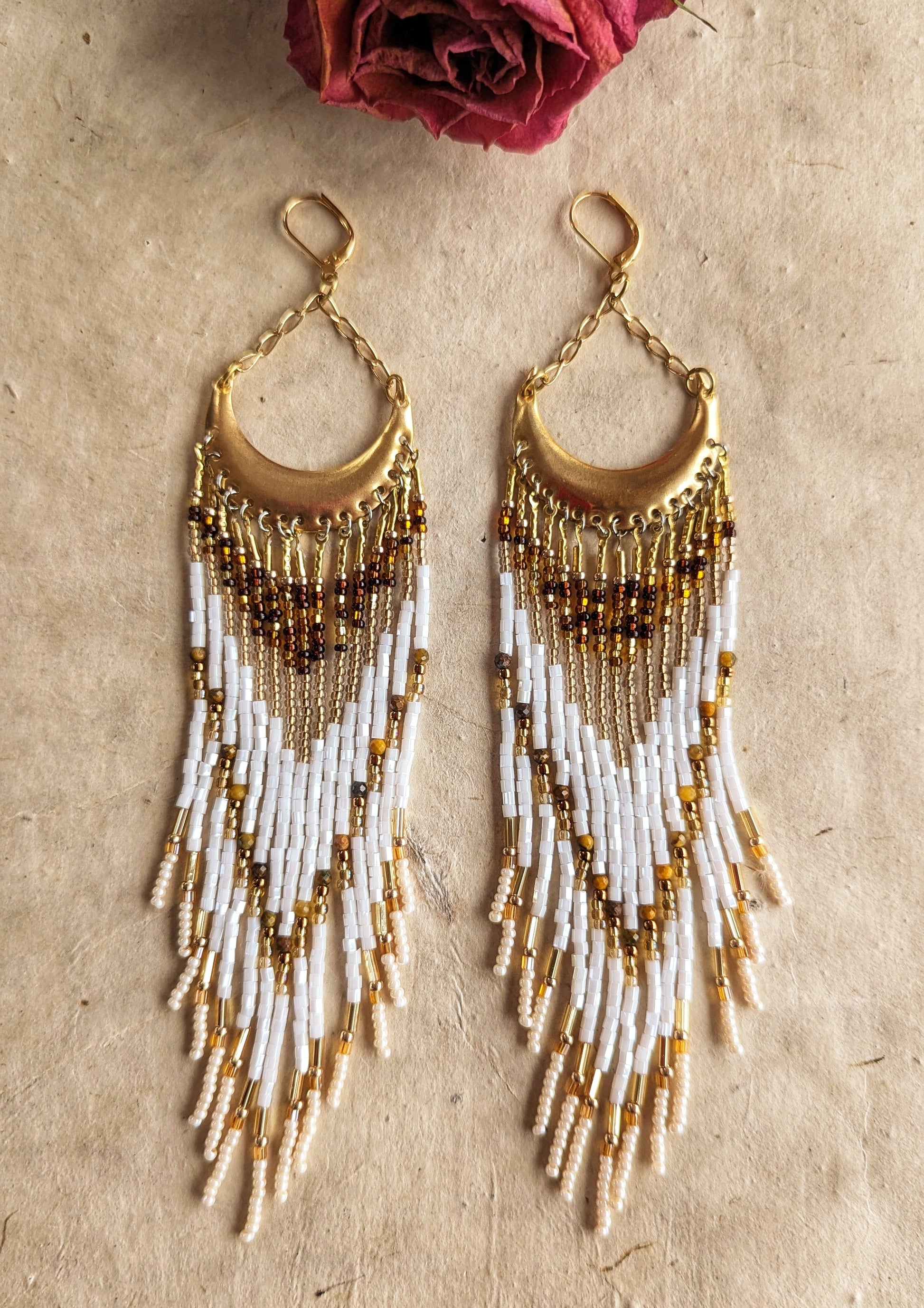 Moon & Milk beaded earrings with crescent brass tops, featuring layered fringes in white, amber, and gold tones, styled on a textured beige background.