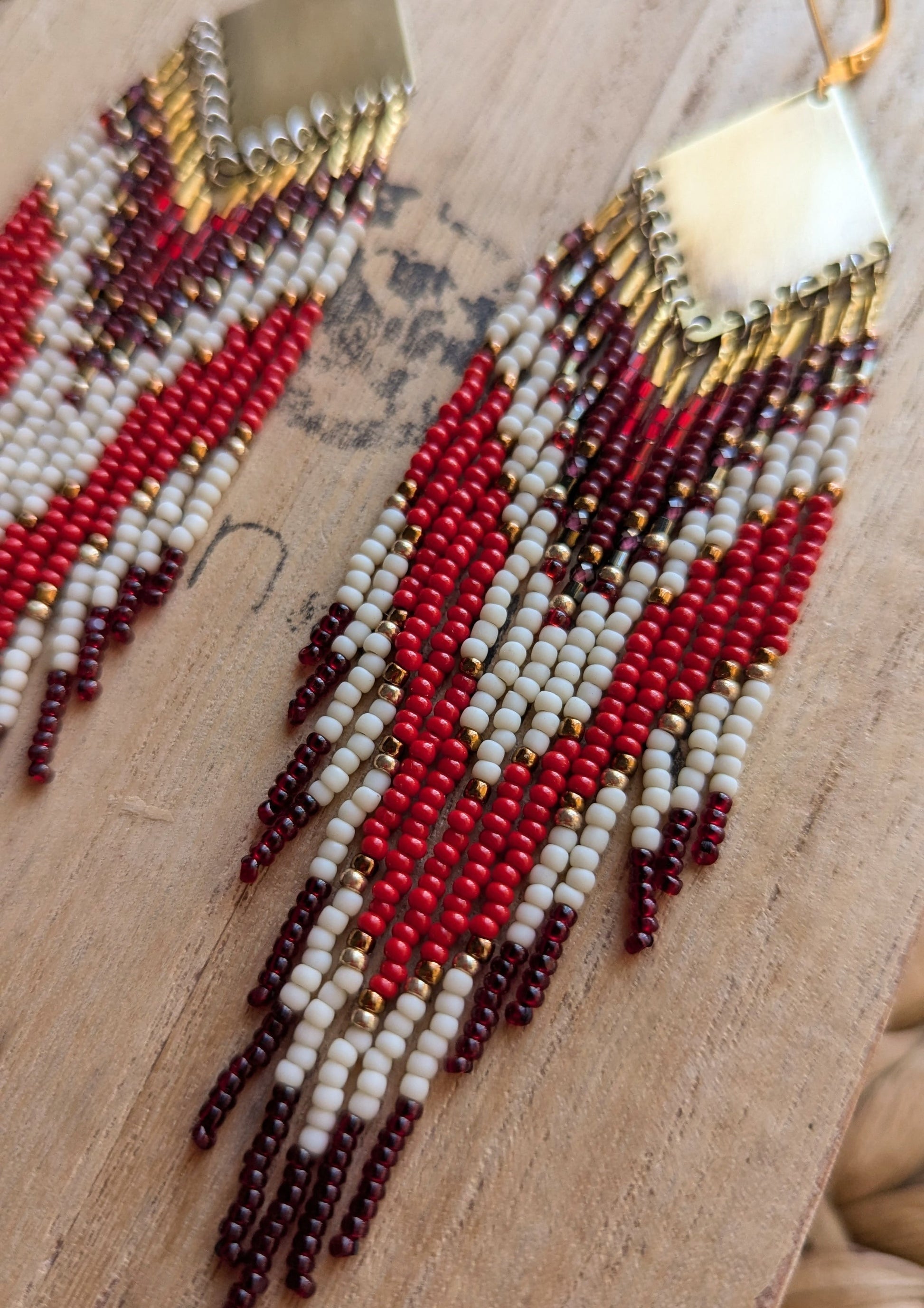 Moon & Milk handmade beaded earrings with burgundy, white, and red beadwork in chevron patterns hanging from brass diamond tops, displayed on a wooden surface.