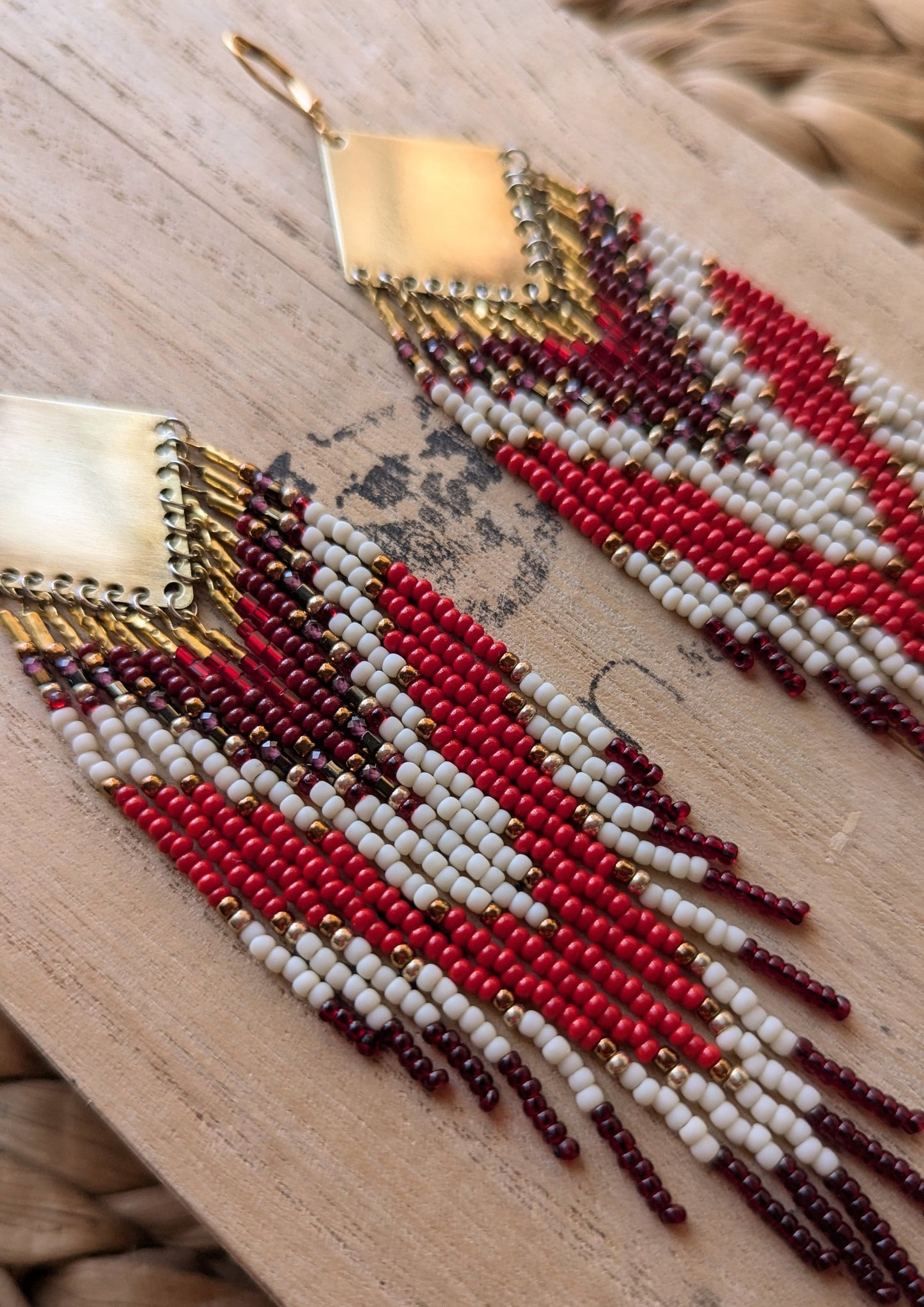Handcrafted beaded earrings featuring a brass top and cascading strands in red, white, and dark maroon, displayed on a wooden surface with a woven background. - Moon & Milk