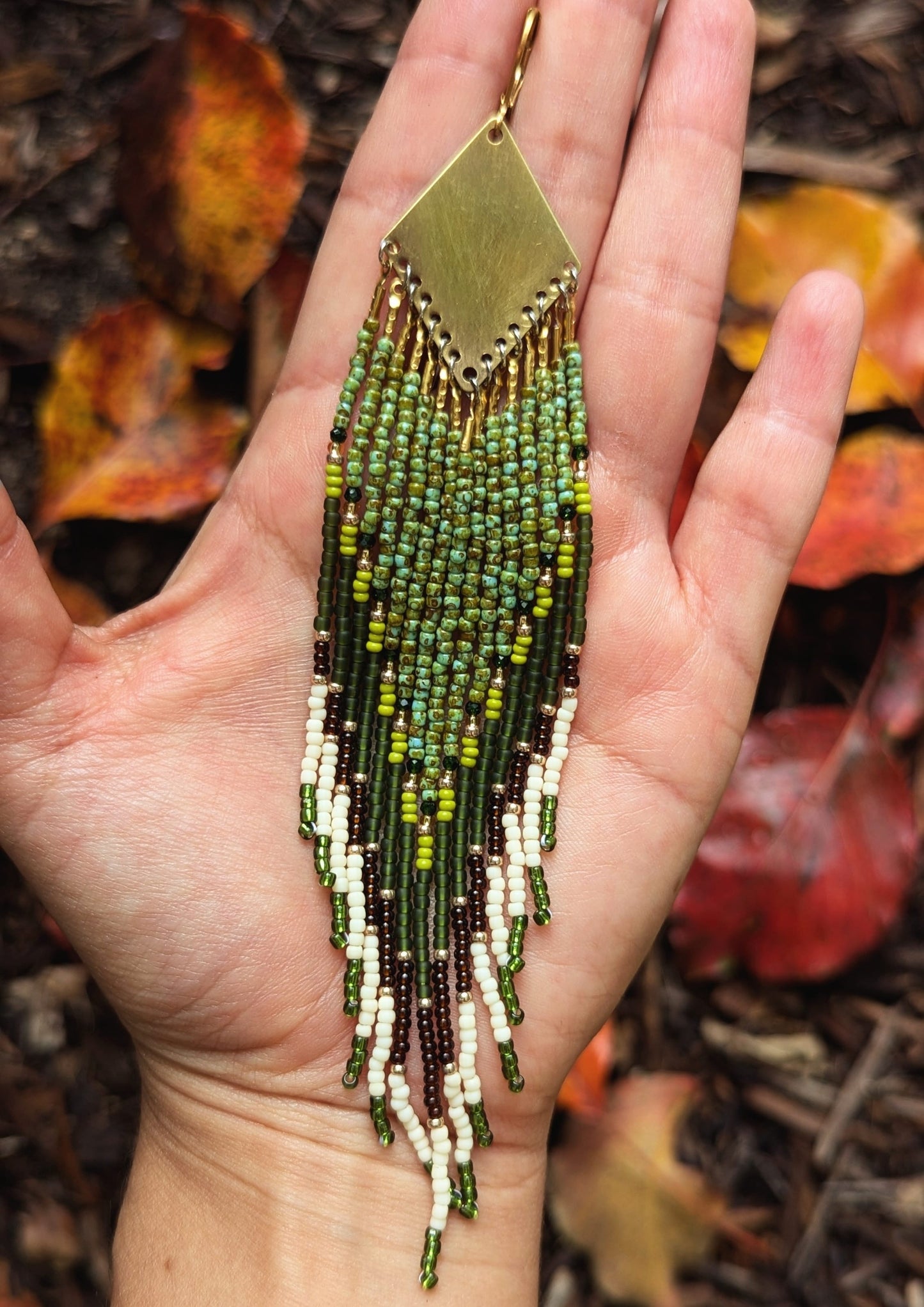 A close-up of a hand holding a beaded earring with a brass top and cascading green, brown, and white beads. Autumn leaves provide a warm background.- Moon & Milk