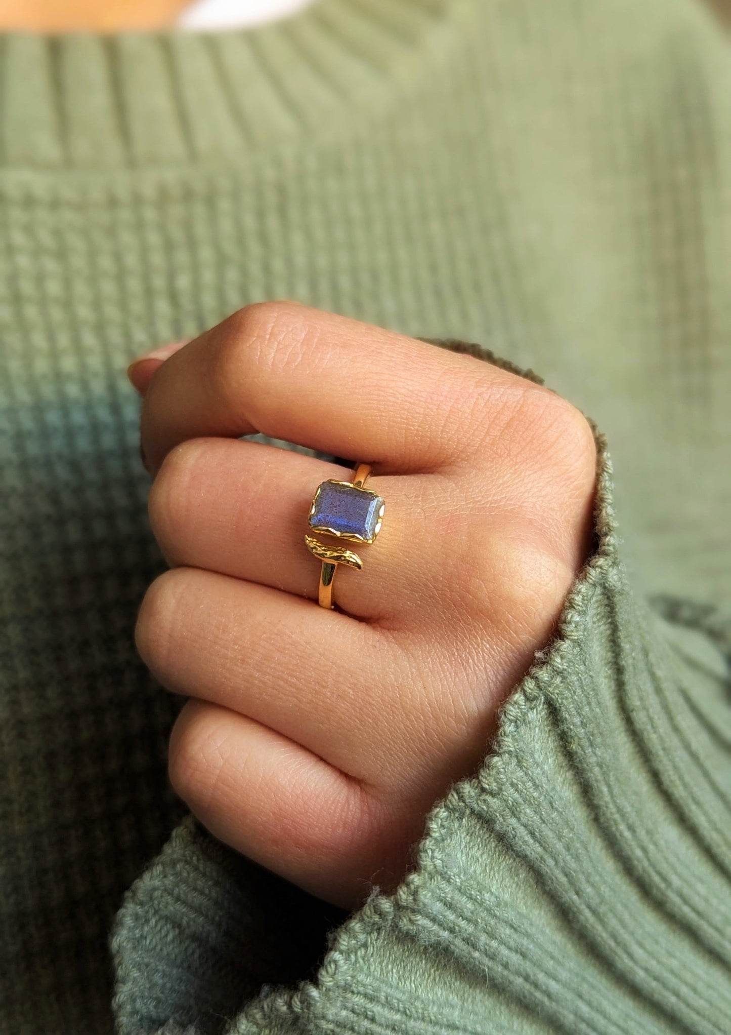 Gold-plated ring with a rectangular labradorite stone and a sculpted branch detail, shown worn on a hand with a cozy green sweater.