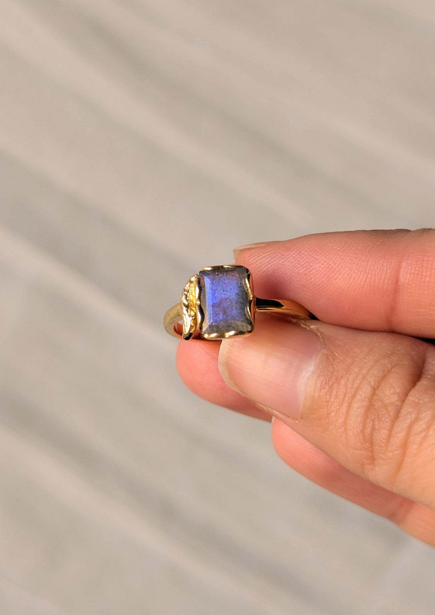 Hand holding a gold ring with a rectangular labradorite stone that catches sunlight, showing its blue flash.