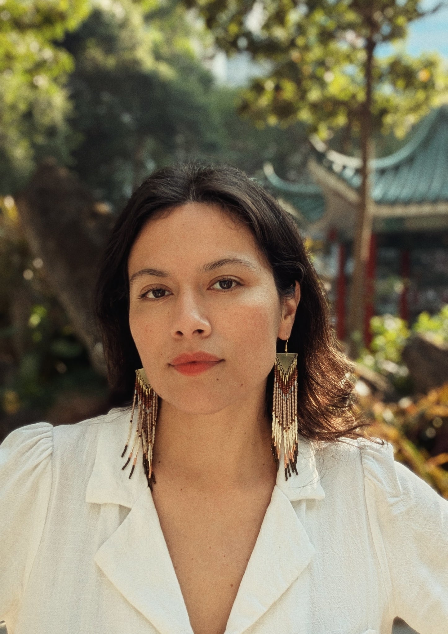 Long handcrafted fringe earrings by Moon & Milk with warm terracotta, beige, and ivory seed beads, finished with brass triangle tops and fine detailing worn by a woman in an Asian garden.