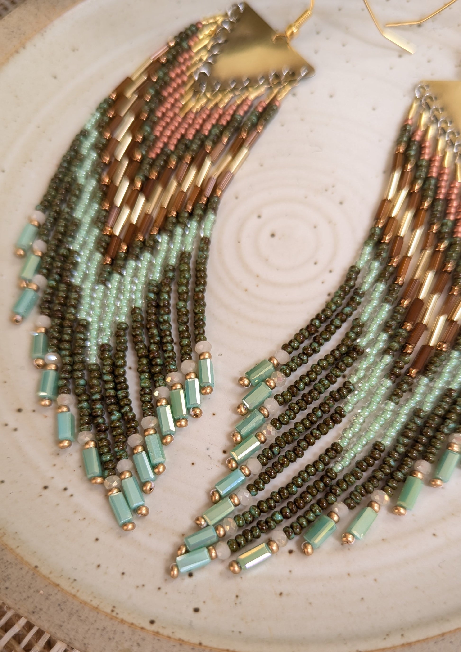Moon & Milk handcrafted fringe earrings with olive green, mint, and bronze seed beads and brass findings, displayed on a ceramic plate in natural light.