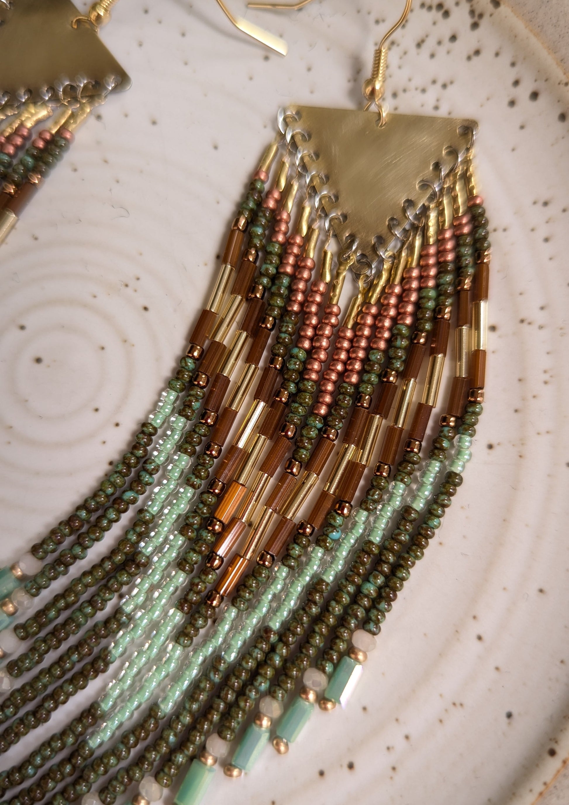 Moon & Milk handcrafted fringe earrings with olive green, mint, and bronze seed beads and brass findings, displayed on a ceramic plate in natural light.