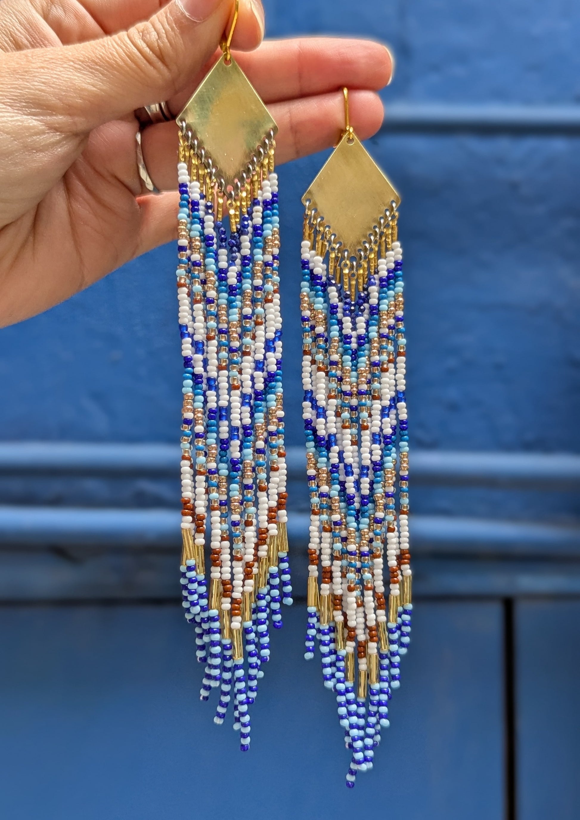 Moon & Milk Blue Origin long native beaded earrings with brass findings, handcrafted with white, cobalt blue, brown, and gold seed beads against a blue wall.