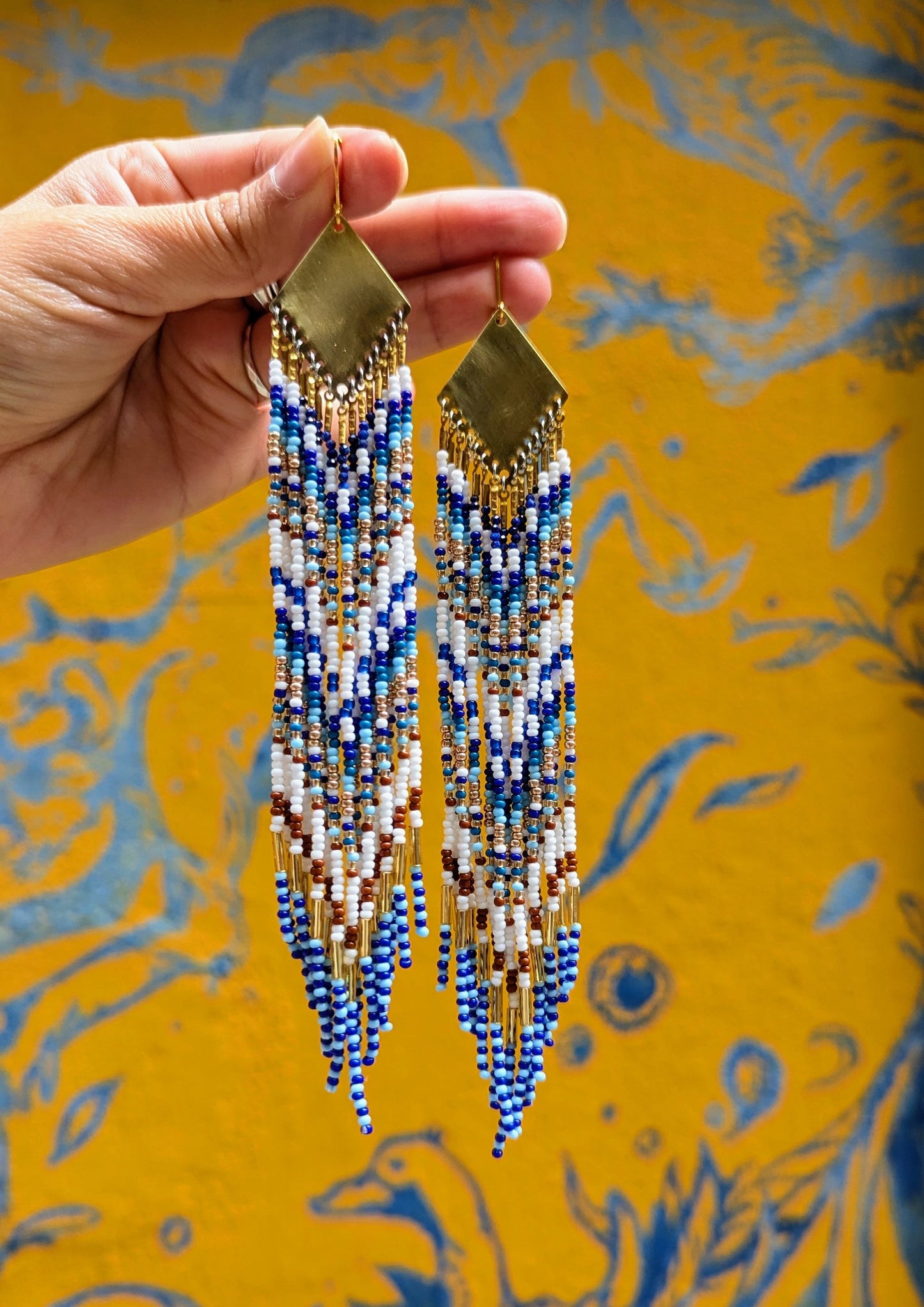 Moon & Milk Blue Origin long native beaded earrings with brass findings, handcrafted with white, cobalt blue, brown, and gold seed beads against a yellow wall.
