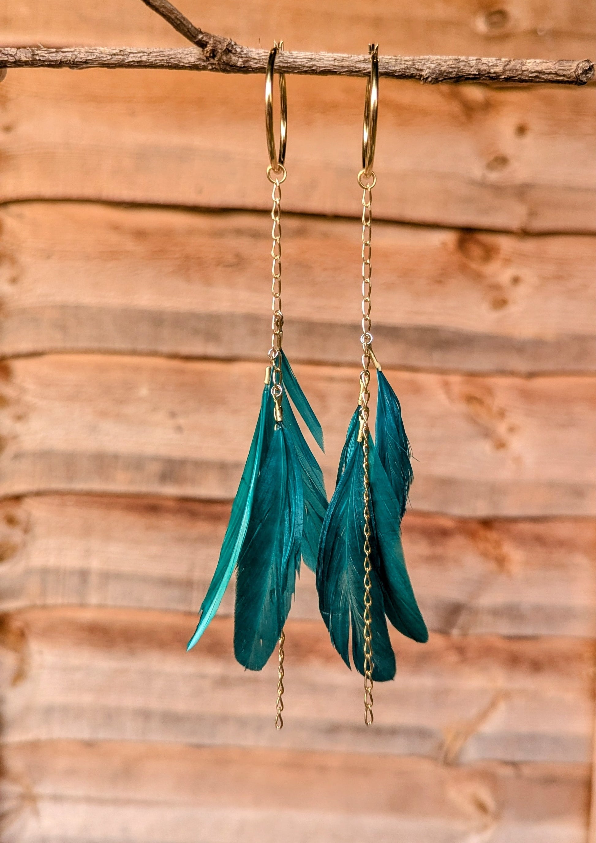 Moon & Milk teal feather earrings with gold chains and hoops, hanging from a branch against a warm wooden background.