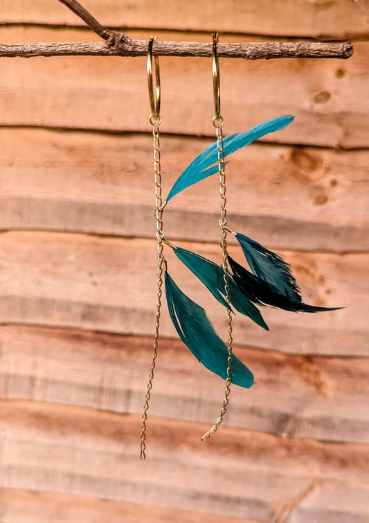 Moon & Milk teal feather earrings with gold chains and hoops, hanging from a branch against a warm wooden background.