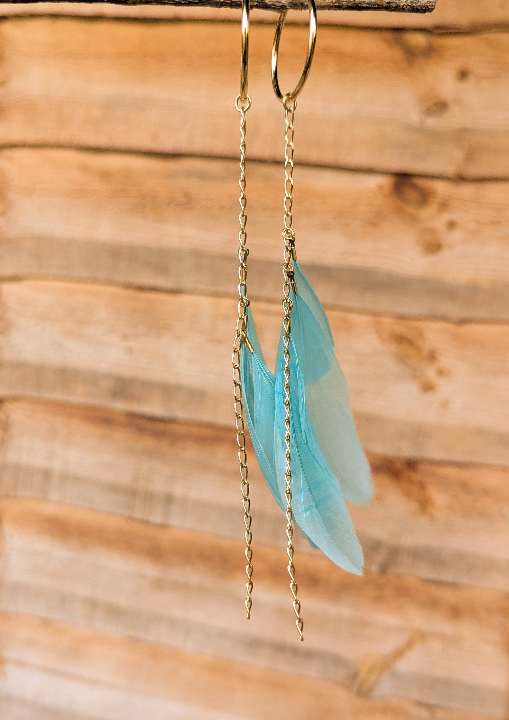 Side view of turquoise feather earrings with long gold chain drops, hanging from gold hoops in front of a wooden background.