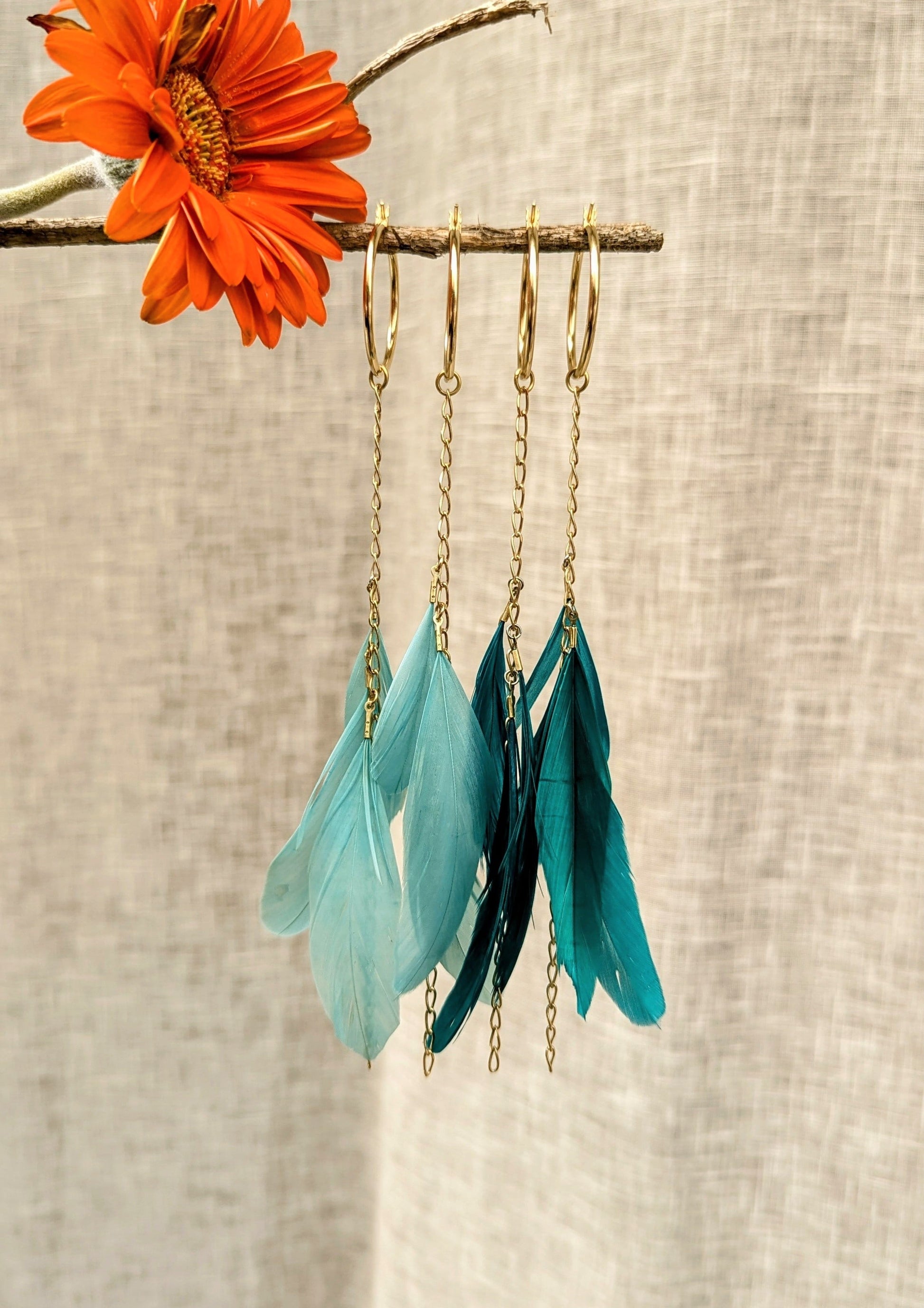 Feather earrings in light turquoise and deep teal tones with gold hoops and dangling chains, displayed on a twig with an orange flower.