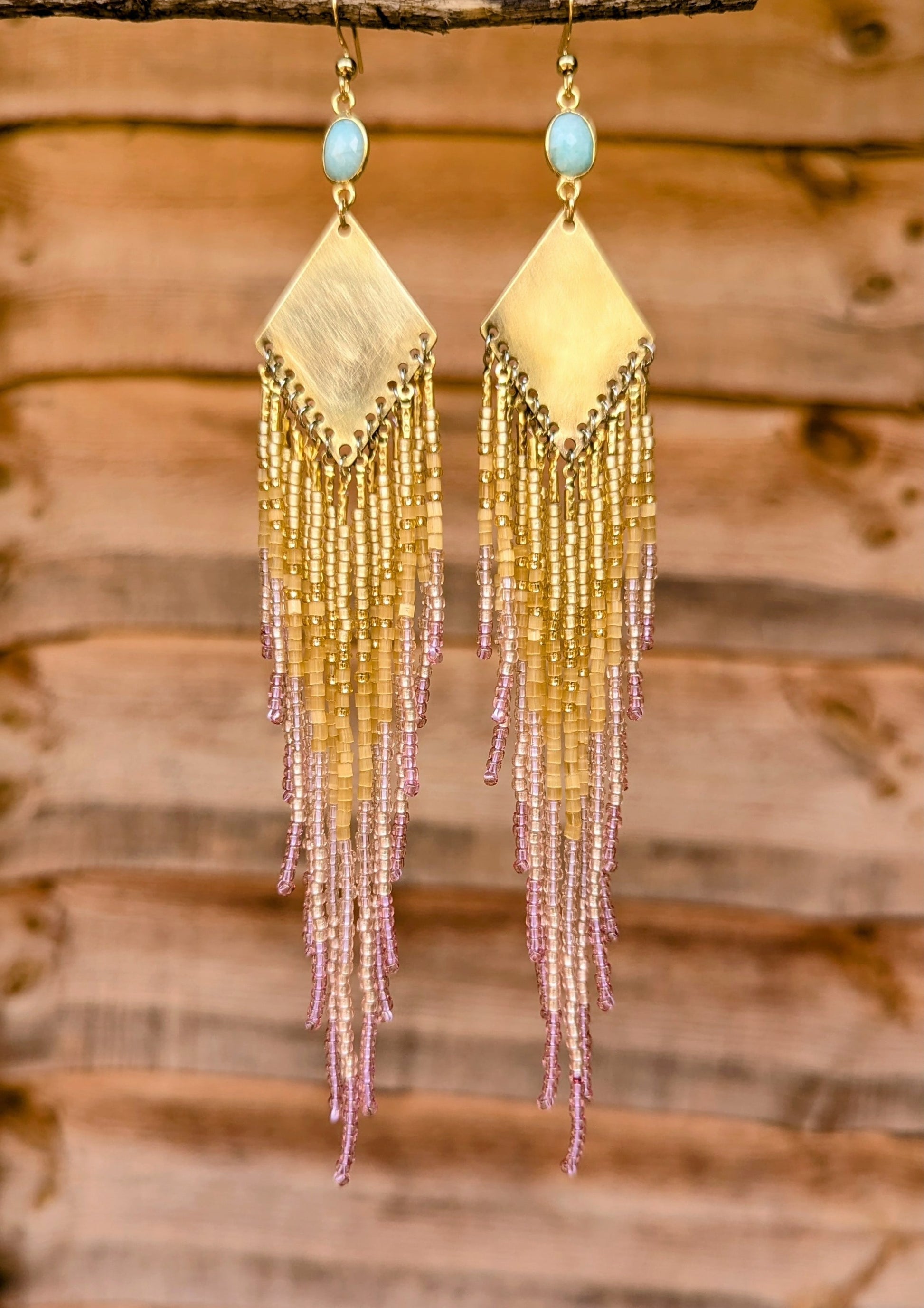 Long beaded earrings by Moon & Milk featuring a brass diamond-shaped base, light pink and gold seed bead fringe, and oval light blue stone studs.