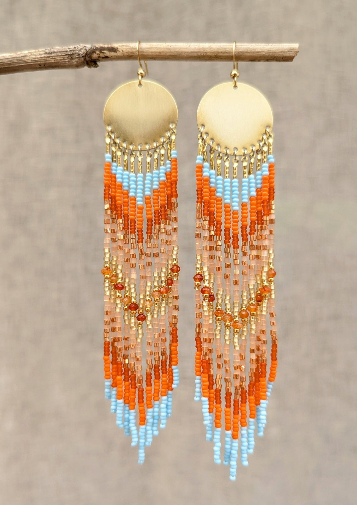 Beaded earrings with bright orange, sky blue, and gold colors hanging on a branch against a neutral background.