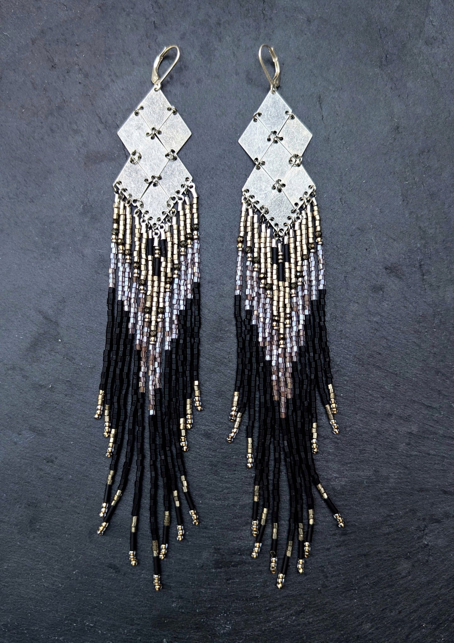 Pair of handcrafted beaded earrings with black, silver, and gold fringes on a dark slate background.