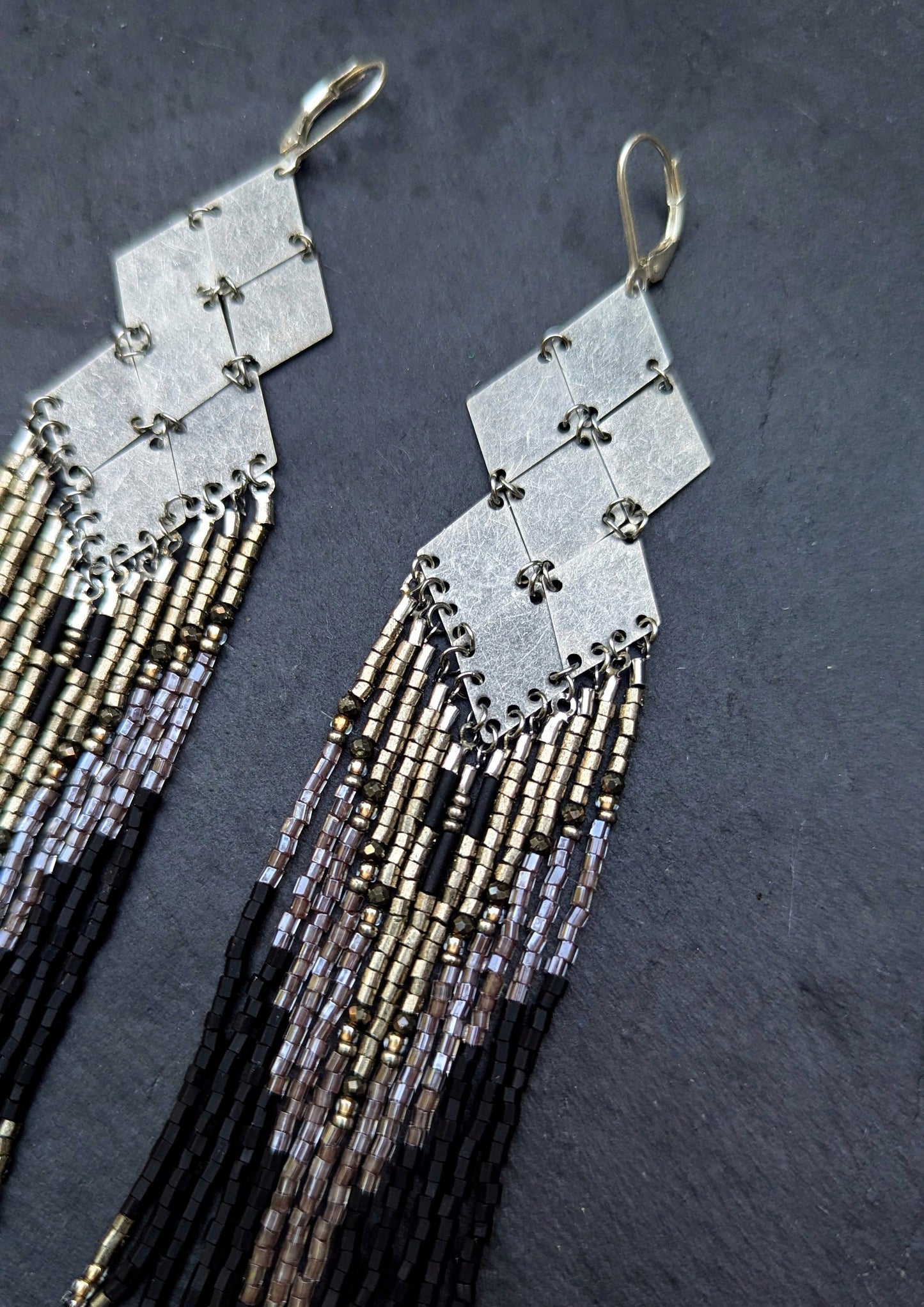 Pair of beaded black earrings with geometric design and fringe details on a dark background.
