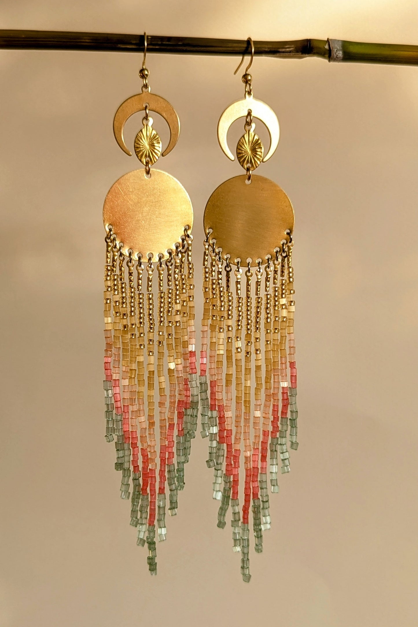 Gold beaded earrings with colorful and sparkly beaded fringes on a beige background.