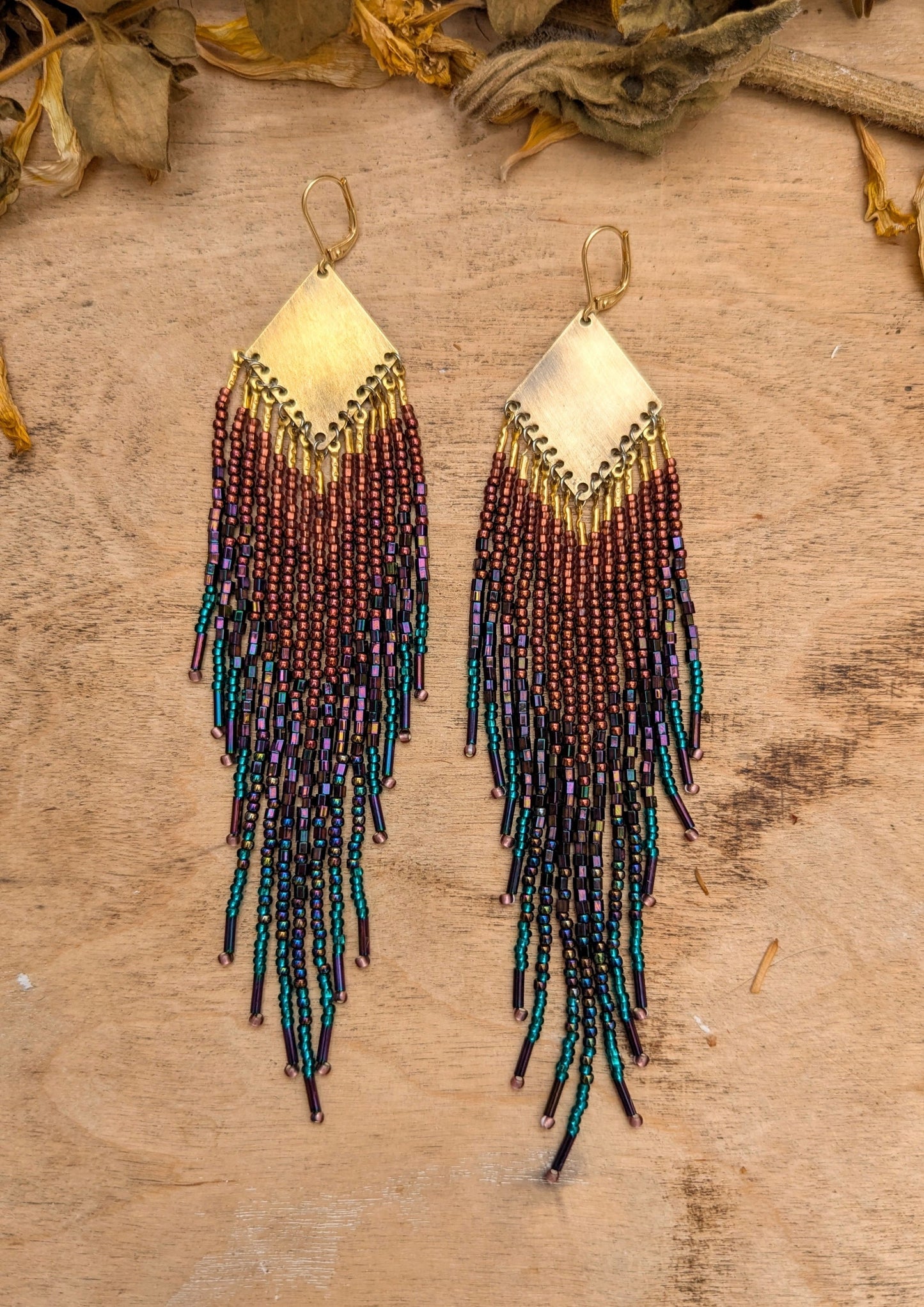 Hand-beaded fringe earrings on a wooden surface with dried leaves.