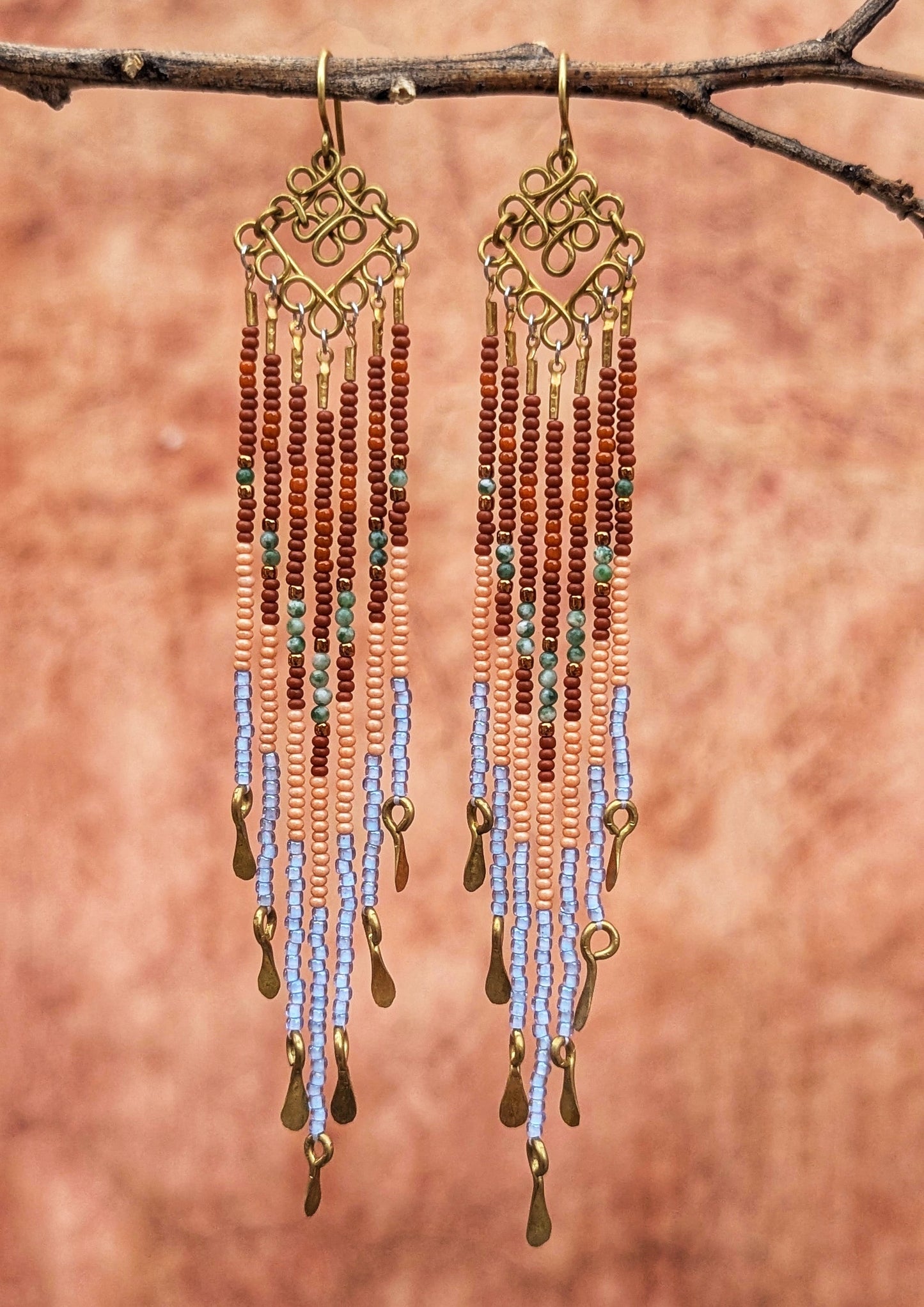 Beaded earrings with gold accents hanging from a branch against a terracotta background