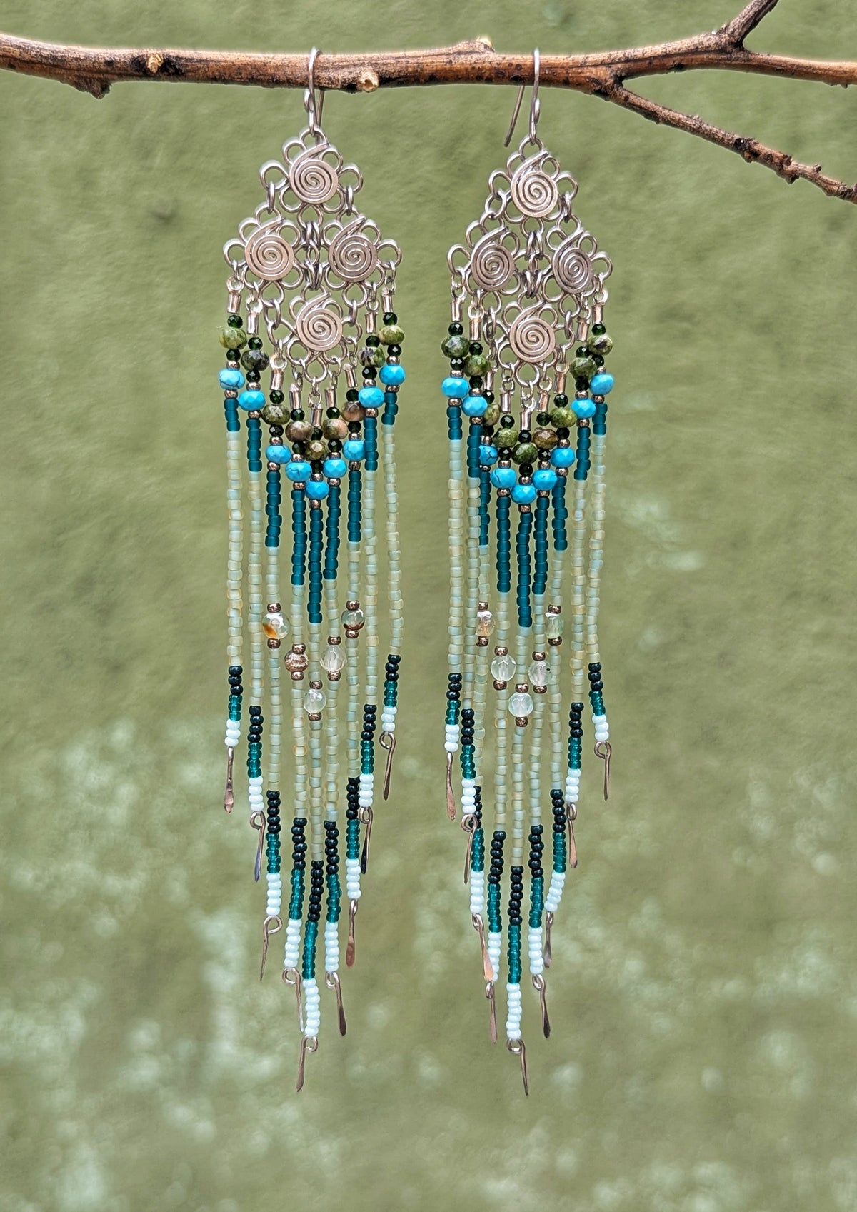 Beaded earrings with blue, green, and silver beads hanging from a branch against a green background.
