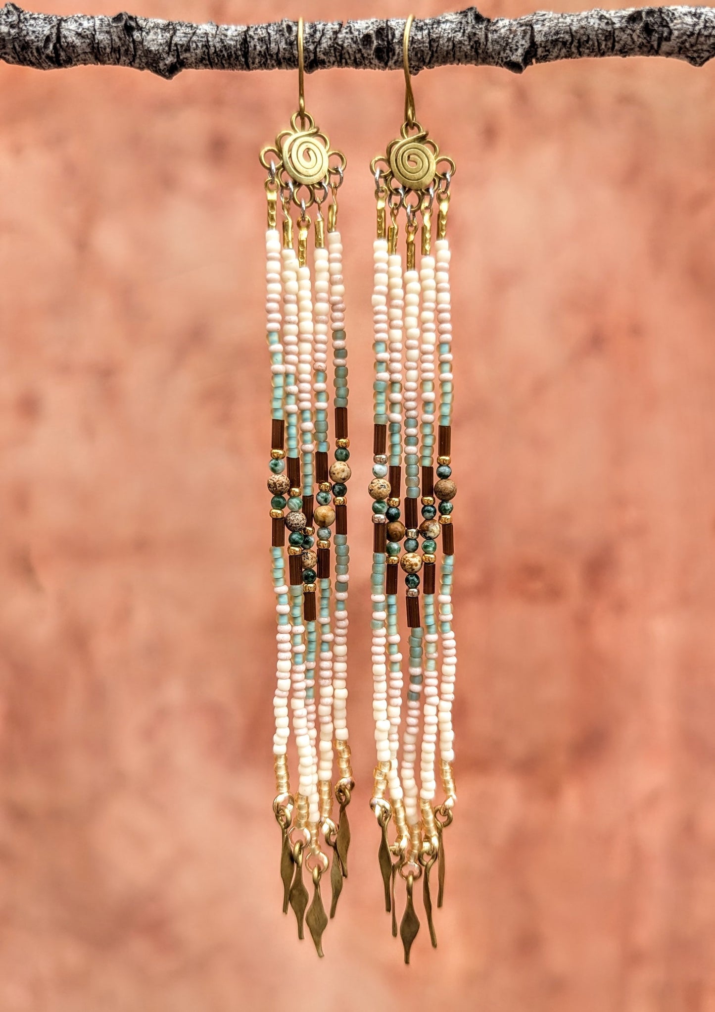 Long beaded earrings with gold accents hanging from a branch against a brown background.