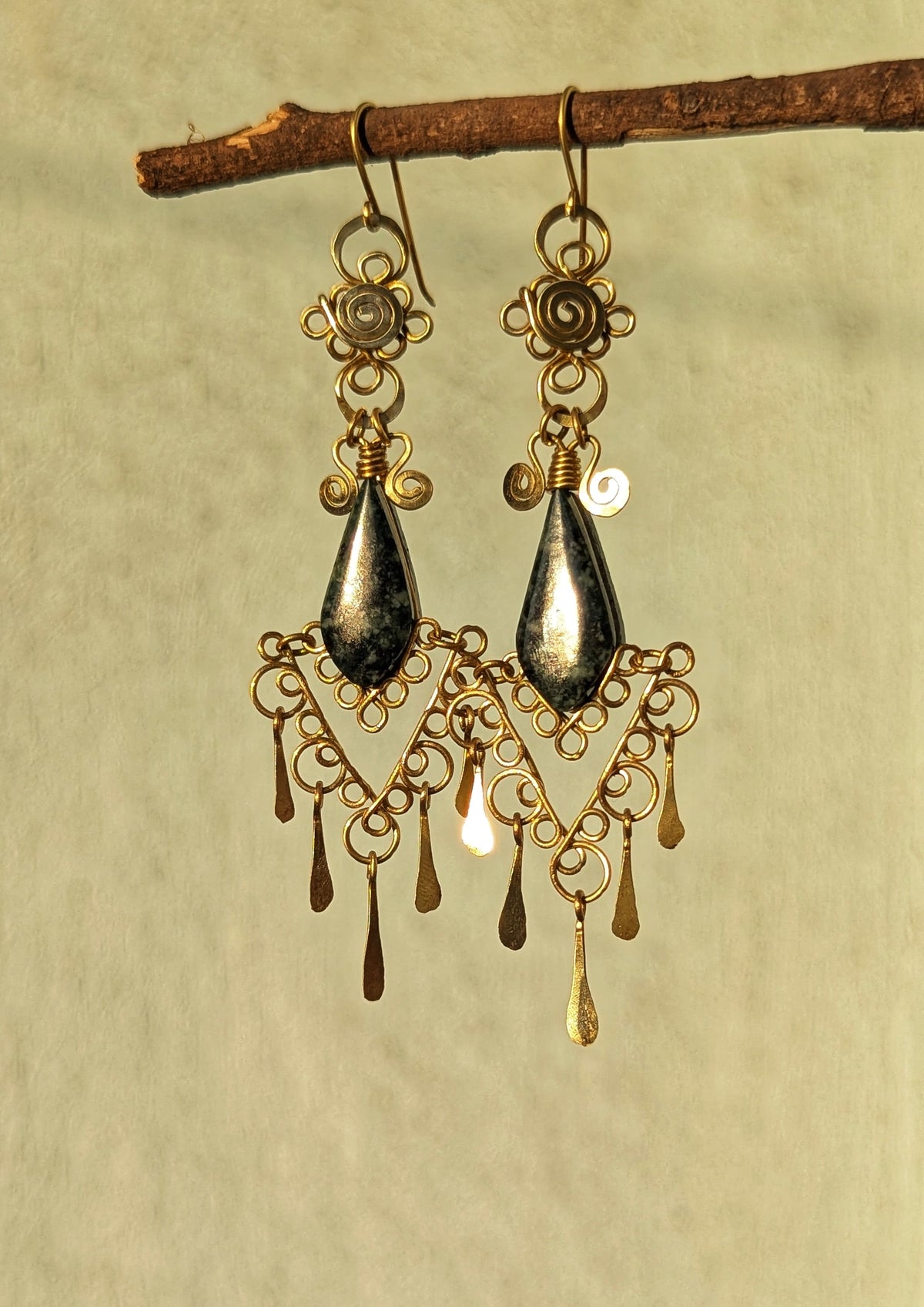 Pair of gold brass earrings hanging from a branch on a beige background.