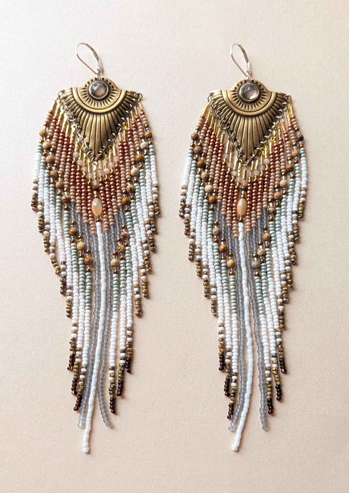 Pair of handmade beaded earrings with gold and white fringes on a beige background.