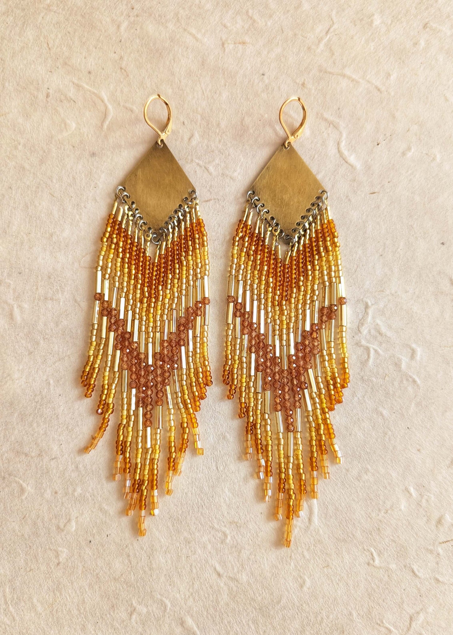 Pair of beaded earrings with gold bugles and hessonite garnet stones, and amber fringes on a beige background.