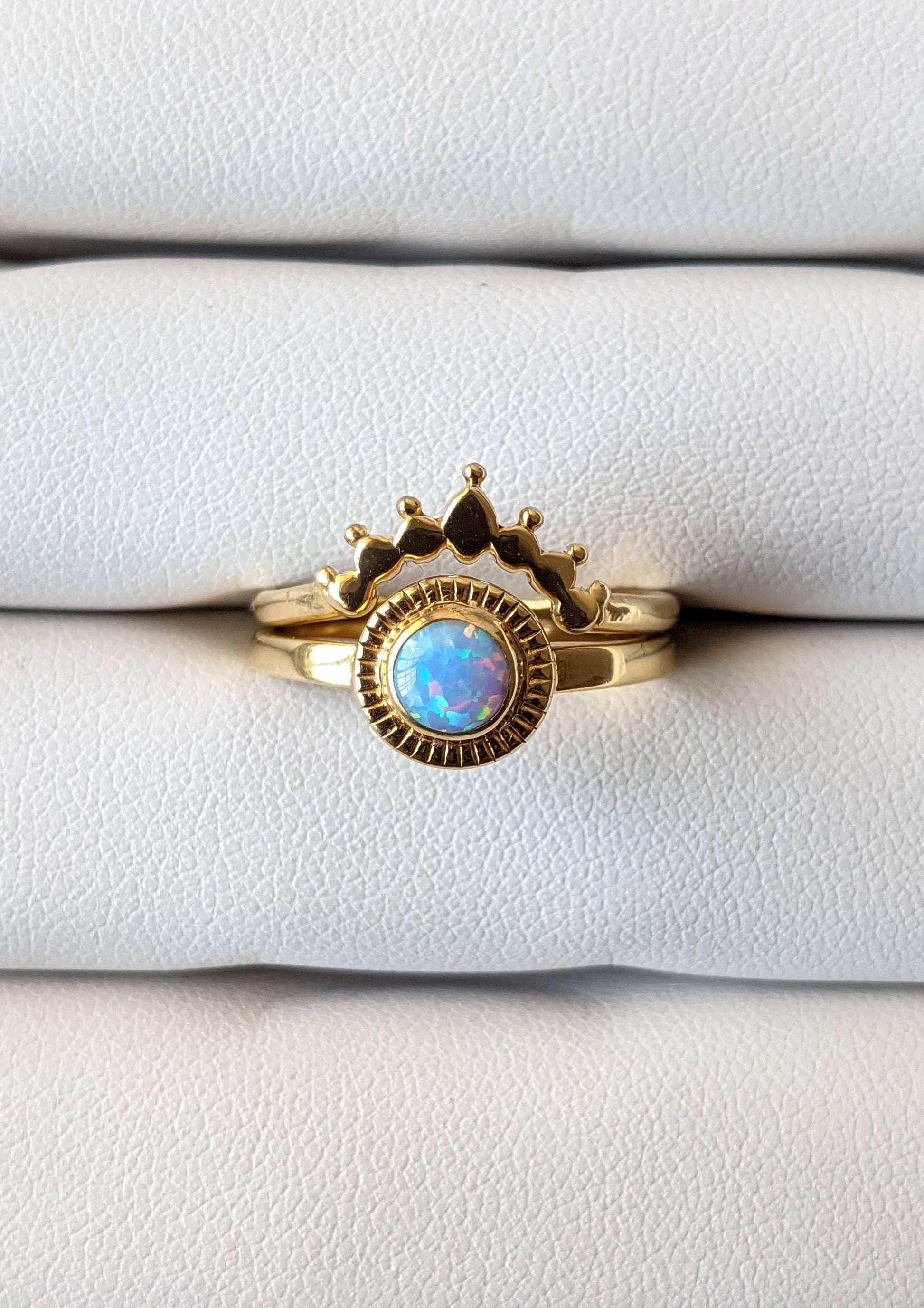 Gold-plated opal ring set featuring a round iridescent stone with sunburst detailing and a crown-shaped stacking band, displayed on a white jewelry holder.