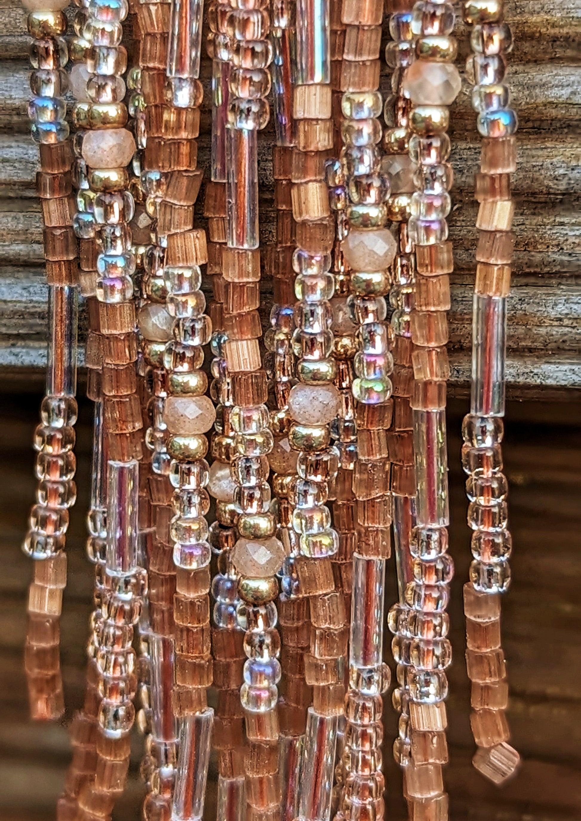 Close-up of Moon & Milk long beaded earrings in shimmering bronze and rose tones with brass findings, laid on a neutral surface in soft sunlight.