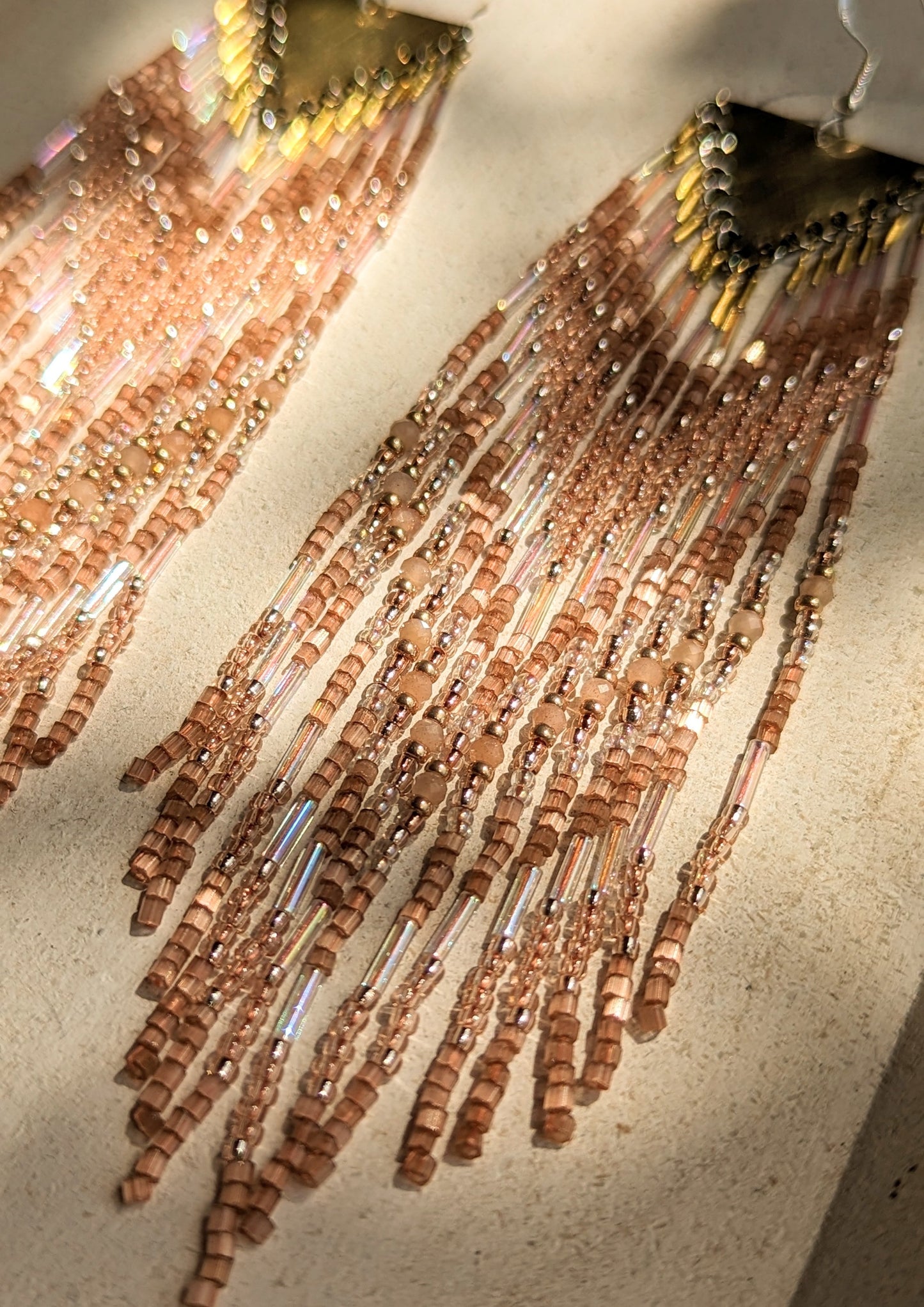 Close-up of Moon & Milk long beaded earrings in shimmering bronze and rose tones with brass findings, laid on a neutral surface in soft sunlight.