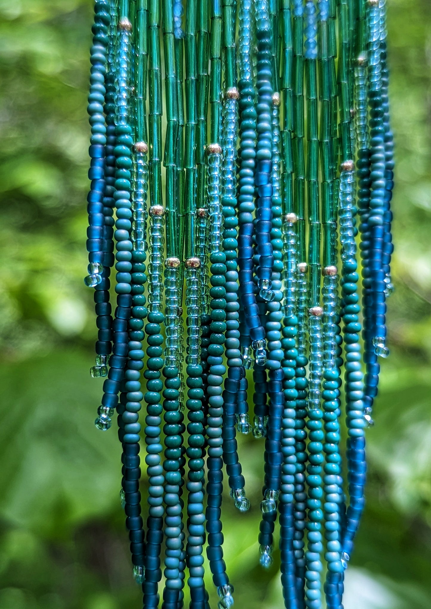 Close-up of long beaded earrings by Moon & Milk with deep green, teal, and blue fringe, brass diamond tops, and handcrafted detail inspired by the Swiss forest.
