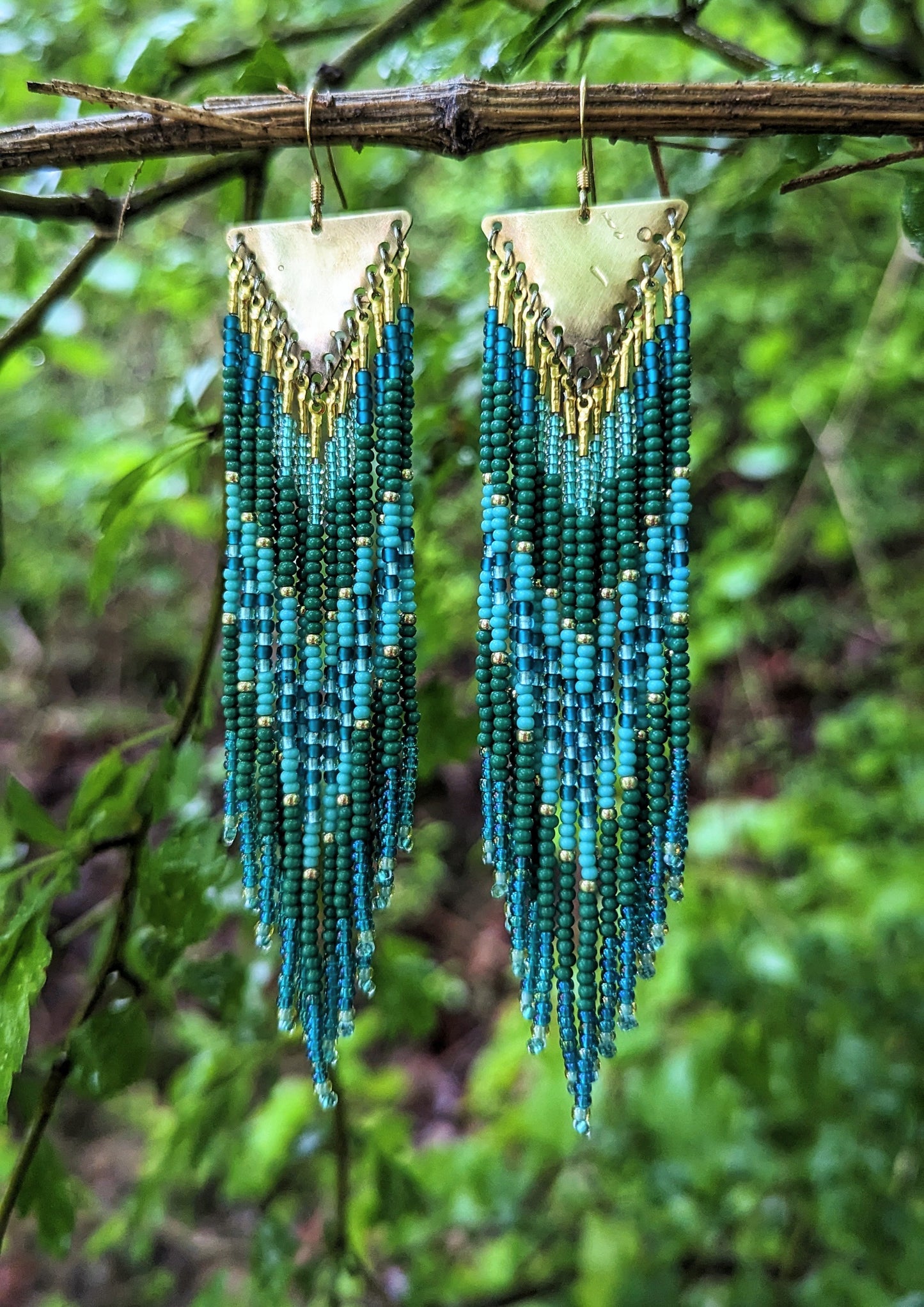 Moon & Milk artisan beaded earrings with layered green, turquoise, and teal beads on brass triangles, hanging in a lush forest setting.