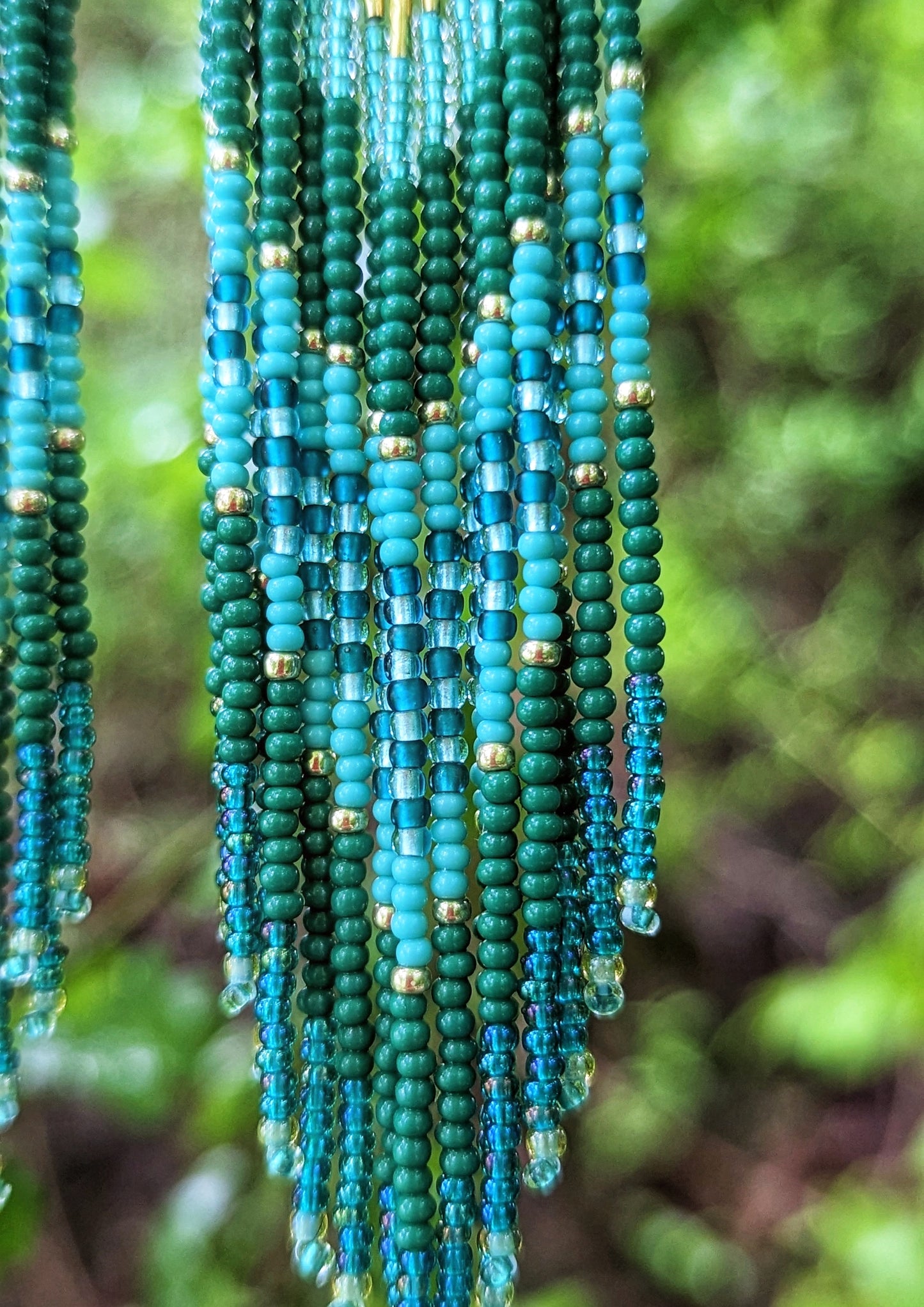 Close-up of Moon & Milk artisan beaded earrings with layered green, turquoise, and teal beads on brass triangles, hanging in a lush forest setting.
