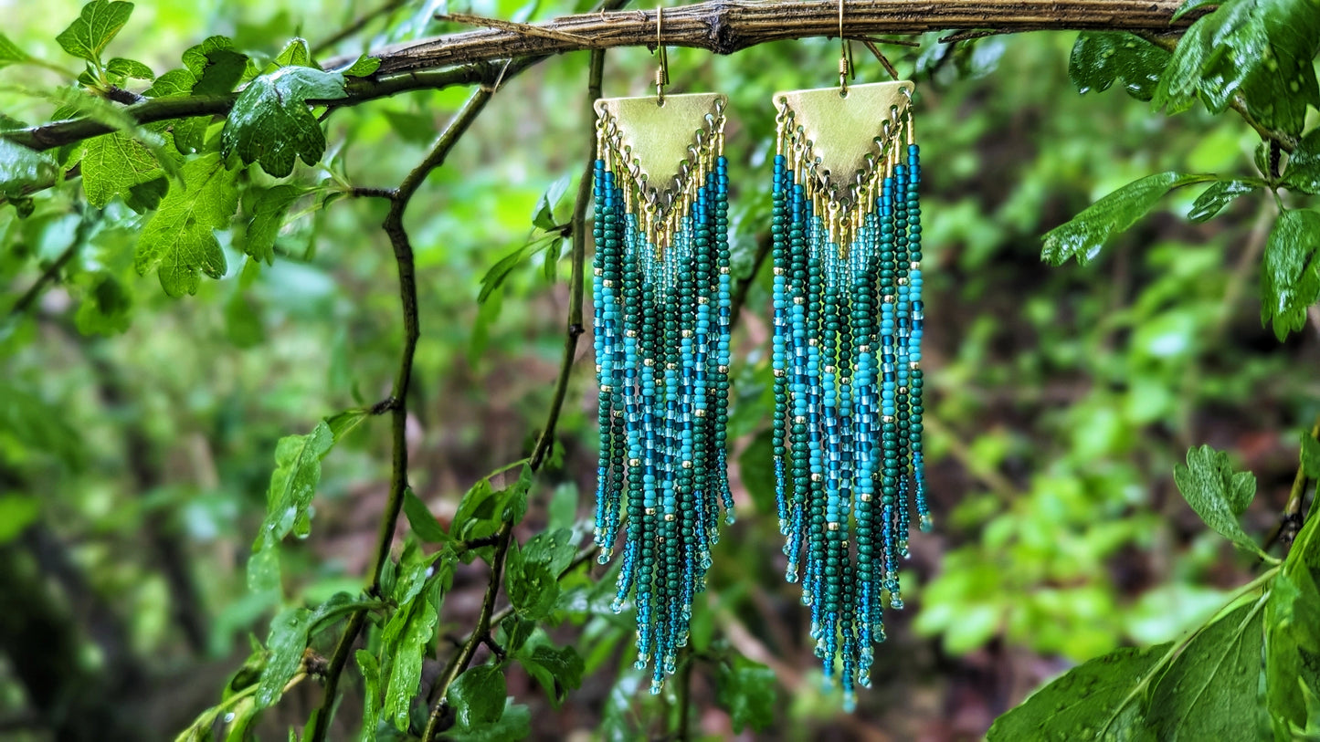 Moon & Milk artisan beaded earrings with layered green, turquoise, and teal beads on brass triangles, hanging in a lush forest setting.