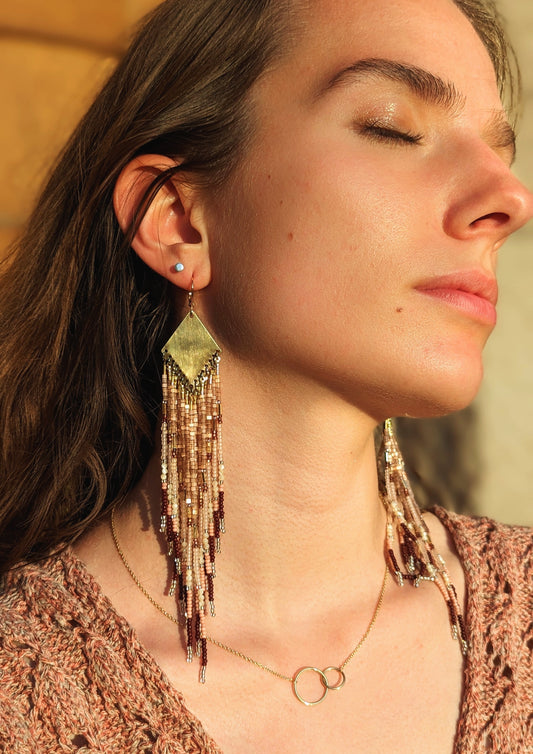 Moon & Milk long native beaded earrings in beige, blush, and bronze tones, worn by a woman in sunlight with a gold double-circle necklace.