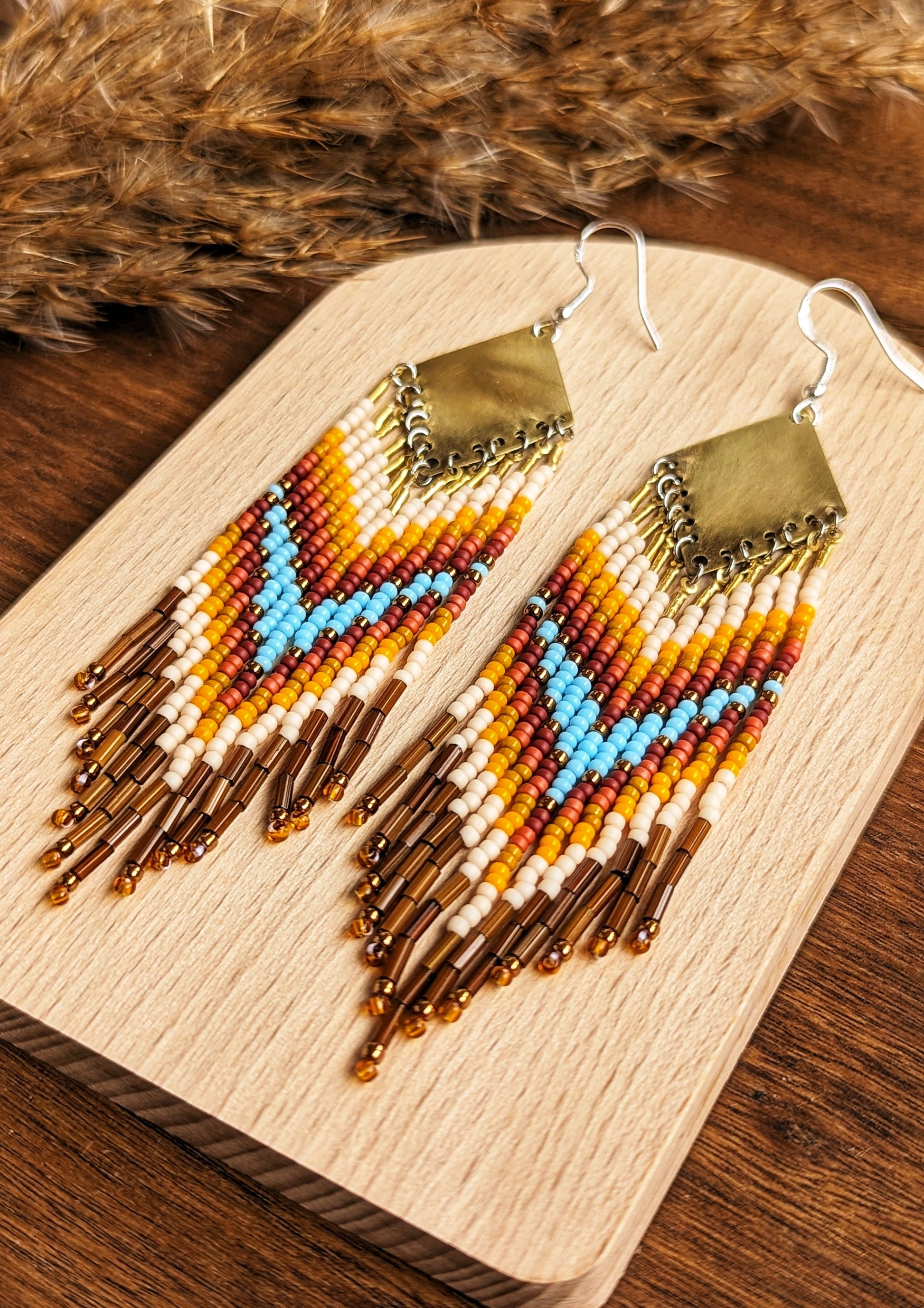 Moon & Milk - Stunning handmade boho beaded earrings featuring a bold chevron pattern with vibrant turquoise, orange, yellow, and brown beads, suspended from a brass diamond-shaped pendant. Perfect statement jewelry for a bohemian-inspired look. Displayed on a wooden board with dried pampas accents for an earthy touch.