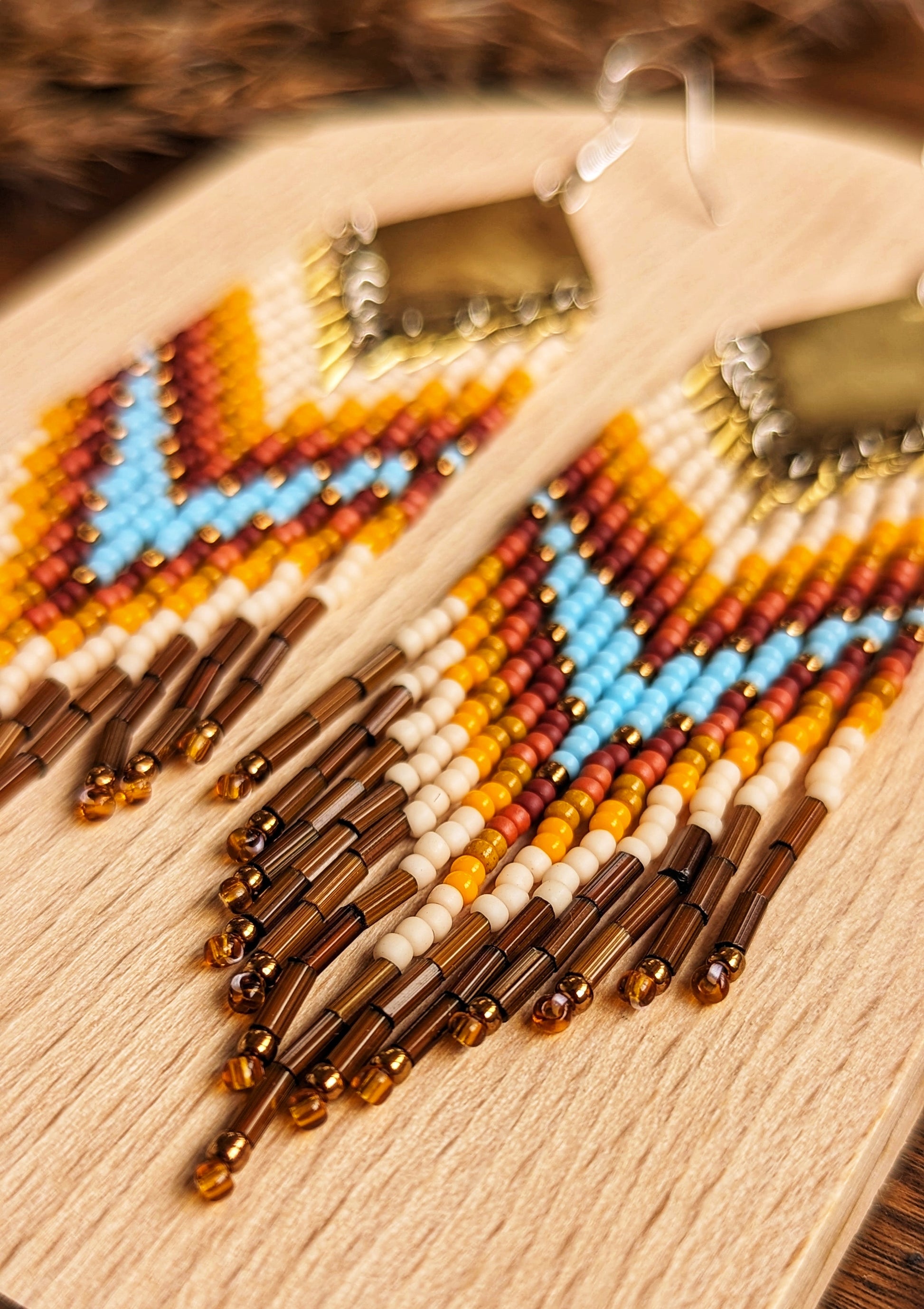 Moon & Milk - Stunning handmade boho beaded earrings featuring a bold chevron pattern with vibrant turquoise, orange, yellow, and brown beads, suspended from a brass diamond-shaped pendant. Perfect statement jewelry for a bohemian-inspired look. Displayed on a wooden board with dried pampas accents for an earthy touch.