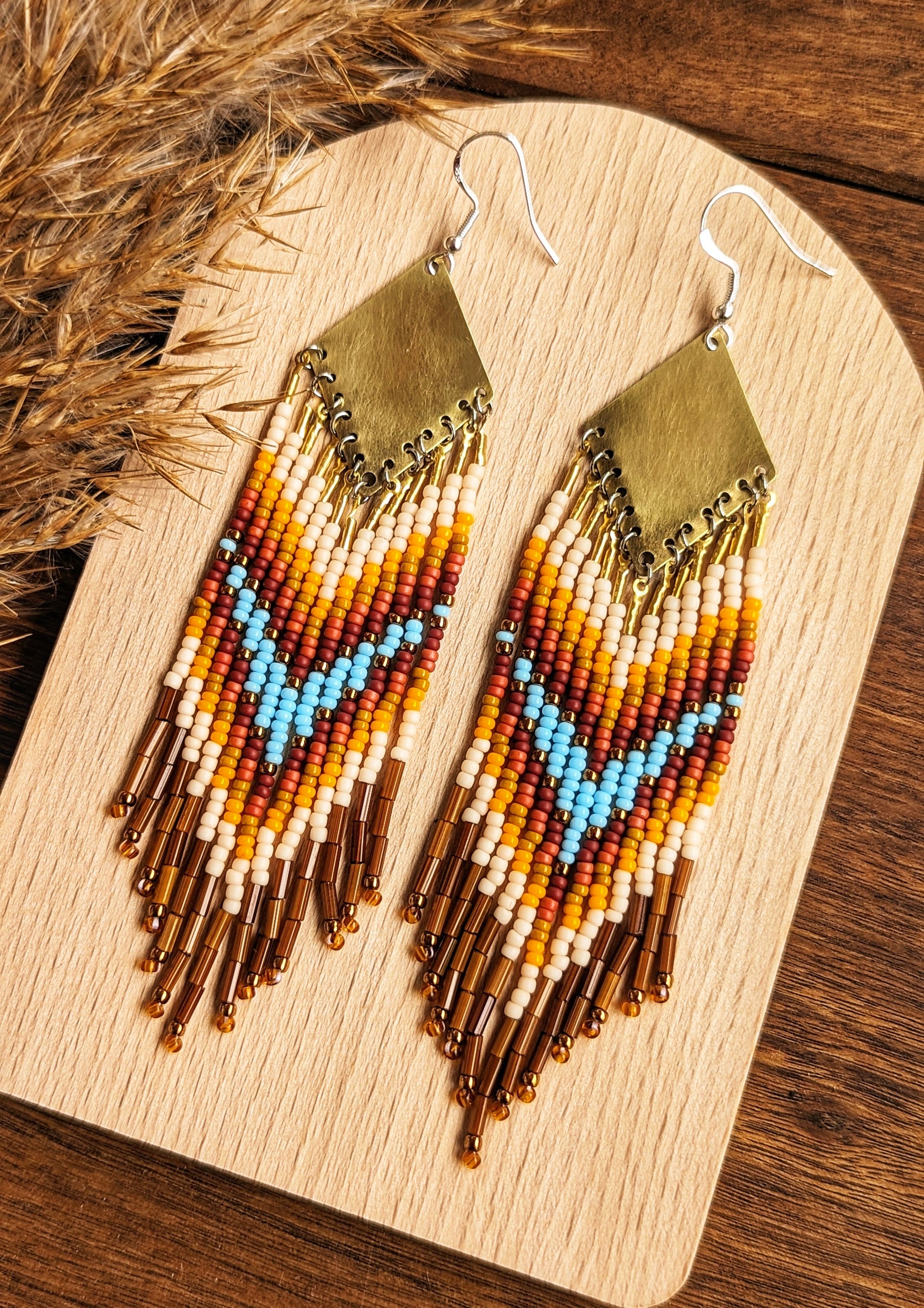 Moon & Milk - Stunning handmade boho beaded earrings featuring a bold chevron pattern with vibrant turquoise, orange, yellow, and brown beads, suspended from a brass diamond-shaped pendant. Perfect statement jewelry for a bohemian-inspired look. Displayed on a wooden board with dried pampas accents for an earthy touch.