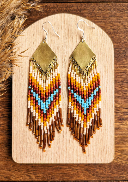 Moon & Milk - Stunning handmade boho beaded earrings featuring a bold chevron pattern with vibrant turquoise, orange, yellow, and brown beads, suspended from a brass diamond-shaped pendant. Perfect statement jewelry for a bohemian-inspired look. Displayed on a wooden board with dried pampas accents for an earthy touch.