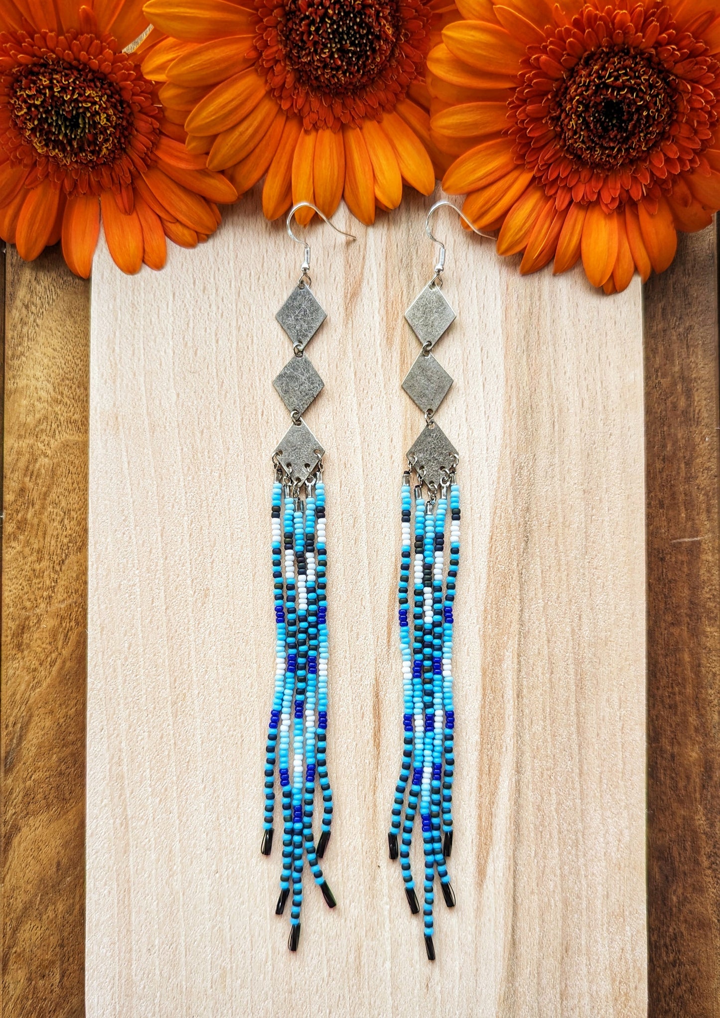 Moon & Milk beaded earrings with silver diamond connectors and long fringe in blue, black, and white tones, displayed on light wood with orange flowers.