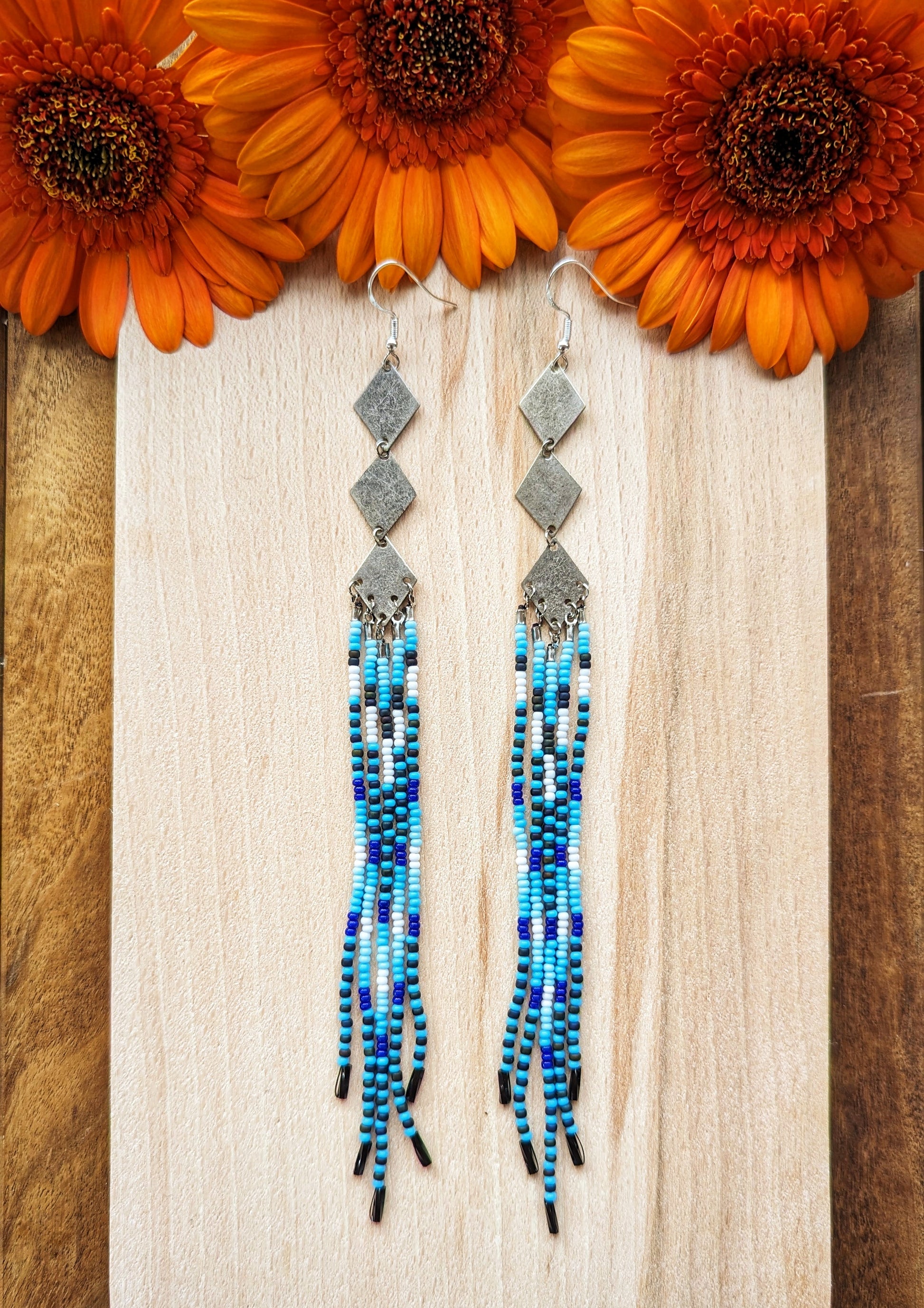 Moon & Milk beaded earrings with silver diamond connectors and long fringe in blue, black, and white tones, displayed on light wood with orange flowers.