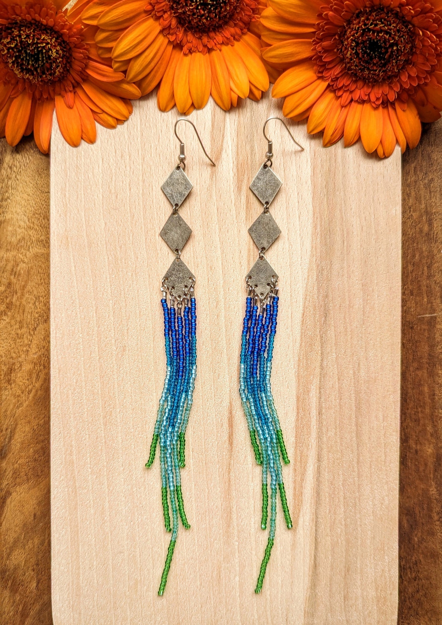 Moon & Milk beaded earrings with silver diamond connectors and long fringe in blue, teal, and green beads, styled with vibrant orange flowers.