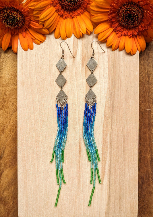 Moon & Milk beaded earrings with silver diamond connectors and long fringe in blue, teal, and green beads, styled with vibrant orange flowers.